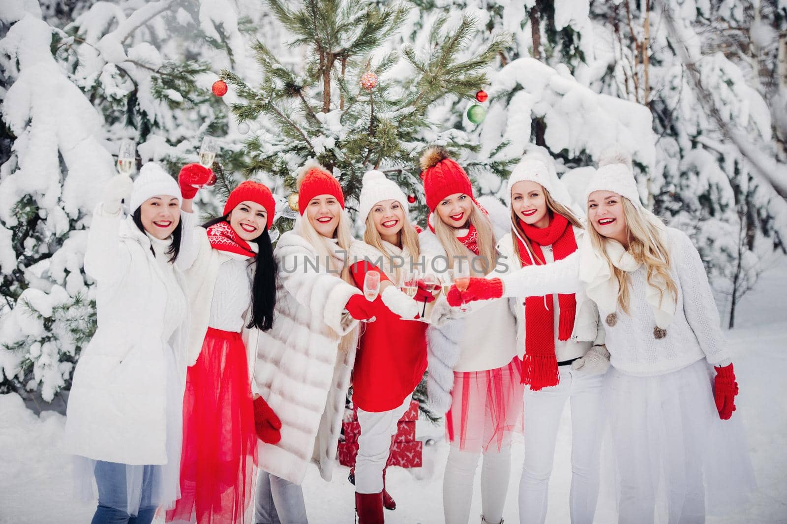 A large group of girls with glasses of champagne in their hands stands in the winter forest.Girls in red and white clothes with new year's drinks in a snow-covered forest. by Lobachad