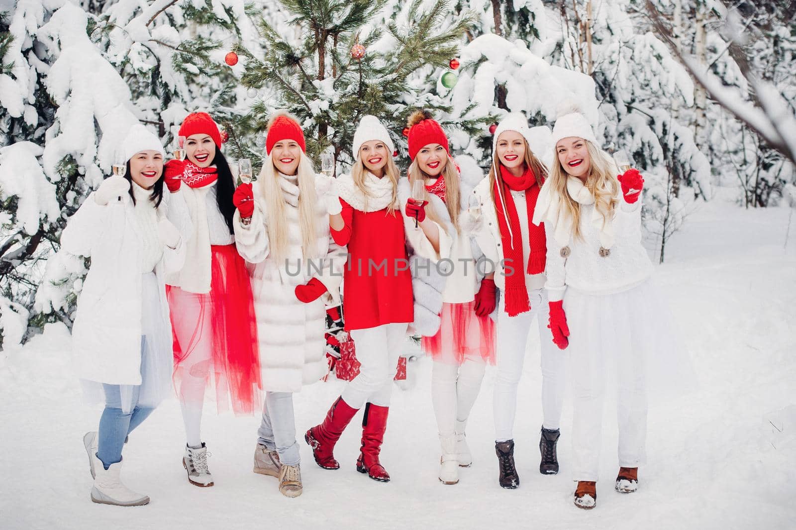 A large group of girls with glasses of champagne in their hands stands in the winter forest.Girls in red and white clothes with new year's drinks in a snow-covered forest