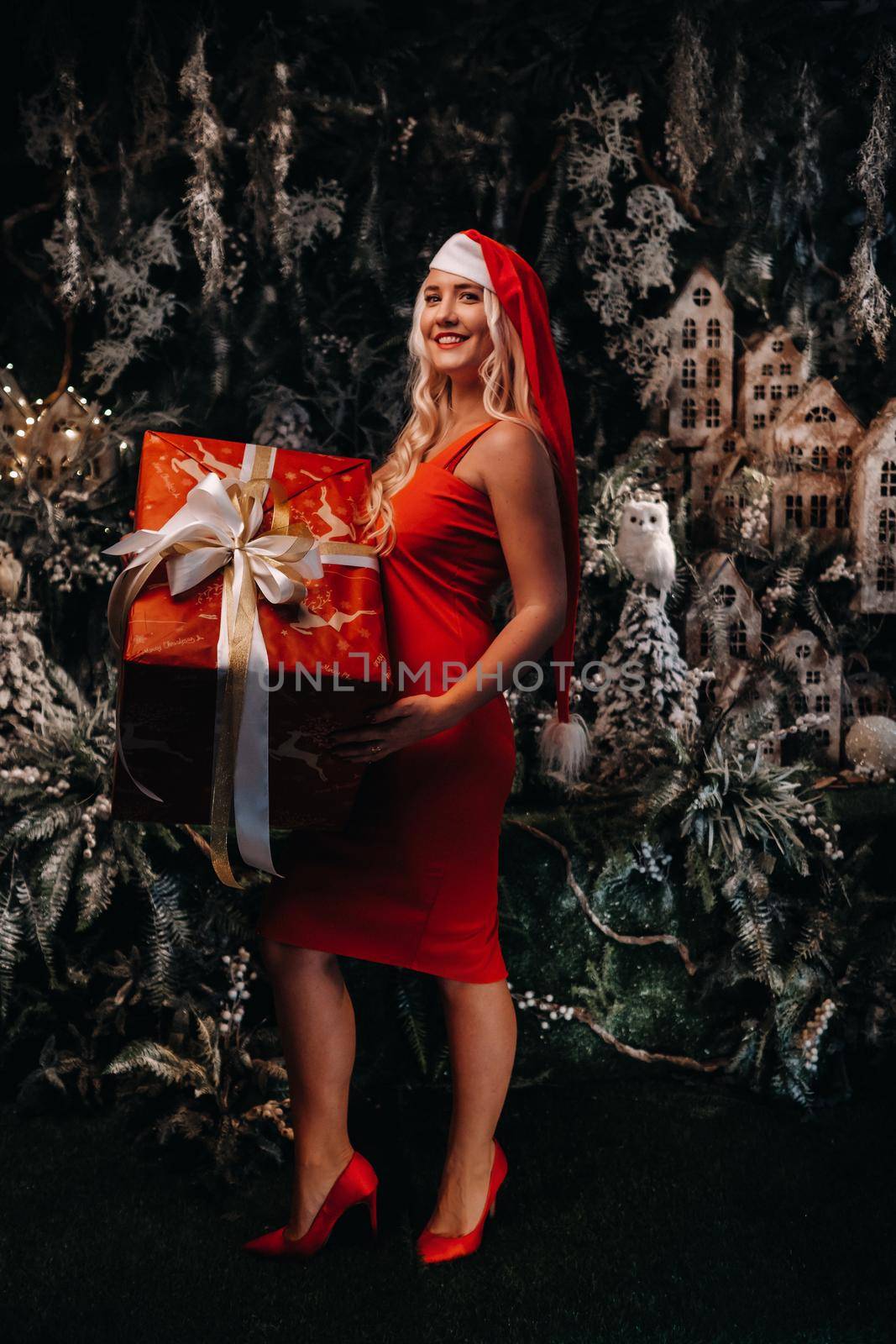 a girl in a Santa hat with a big Christmas gift in her hands on a fabulous Christmas background.A smiling woman in a red dress on the background of Christmas trees and small houses.