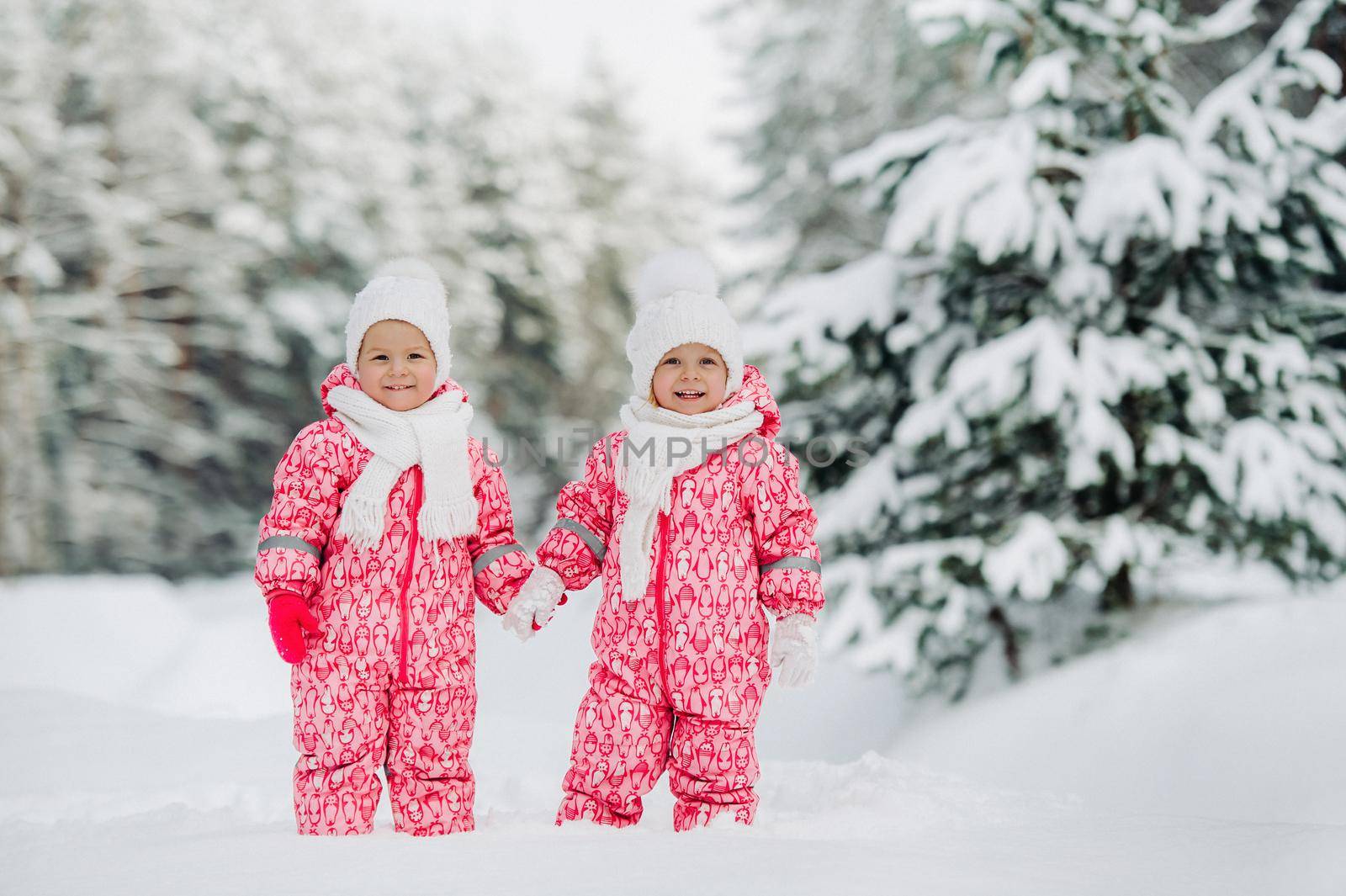 Two little twin girls in red suits stand in a snowy winter forest.