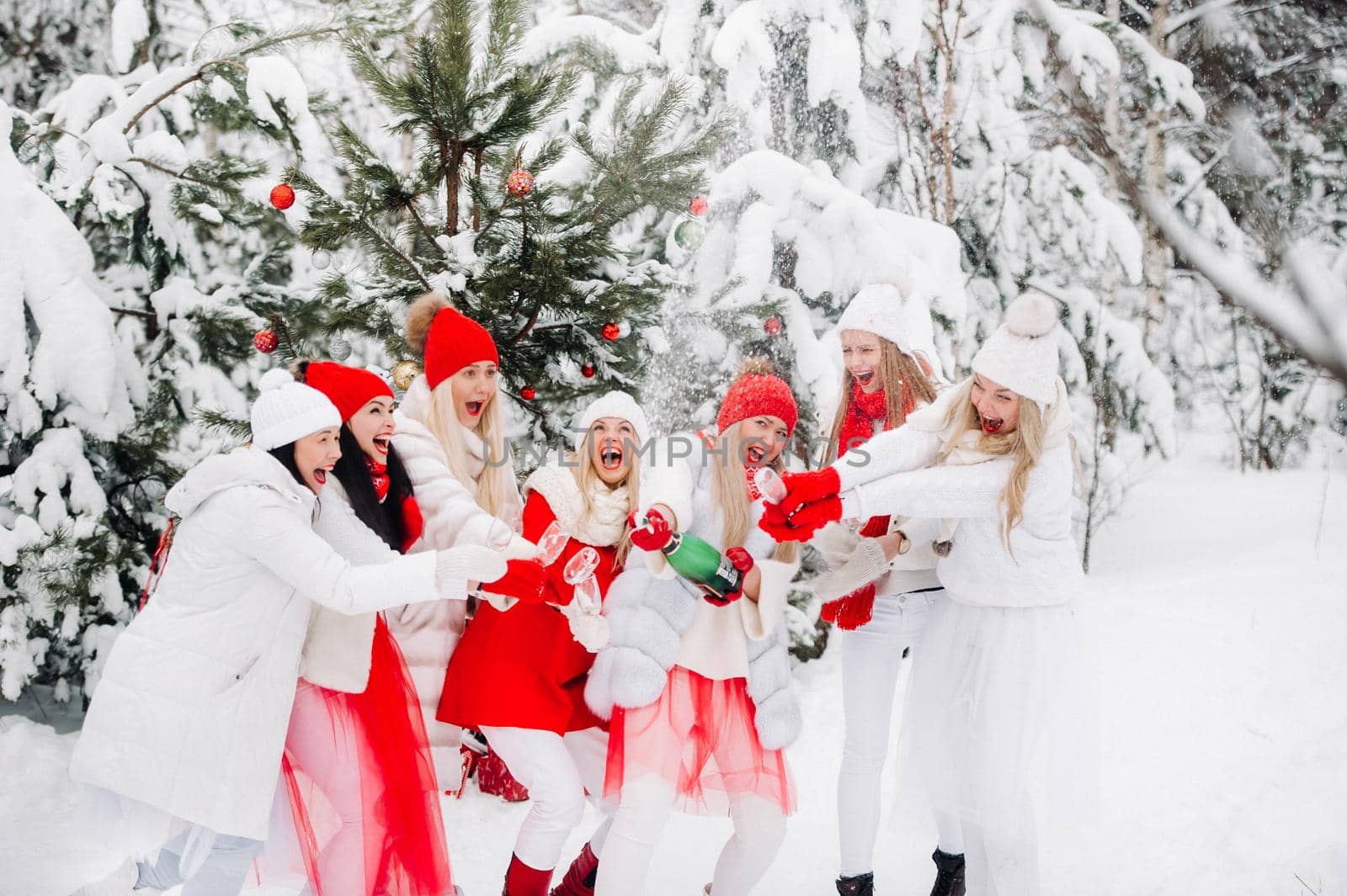 A large group of girls with glasses of champagne in their hands stands in the winter forest.Girls in red and white clothes with new year's drinks in a snow-covered forest. by Lobachad