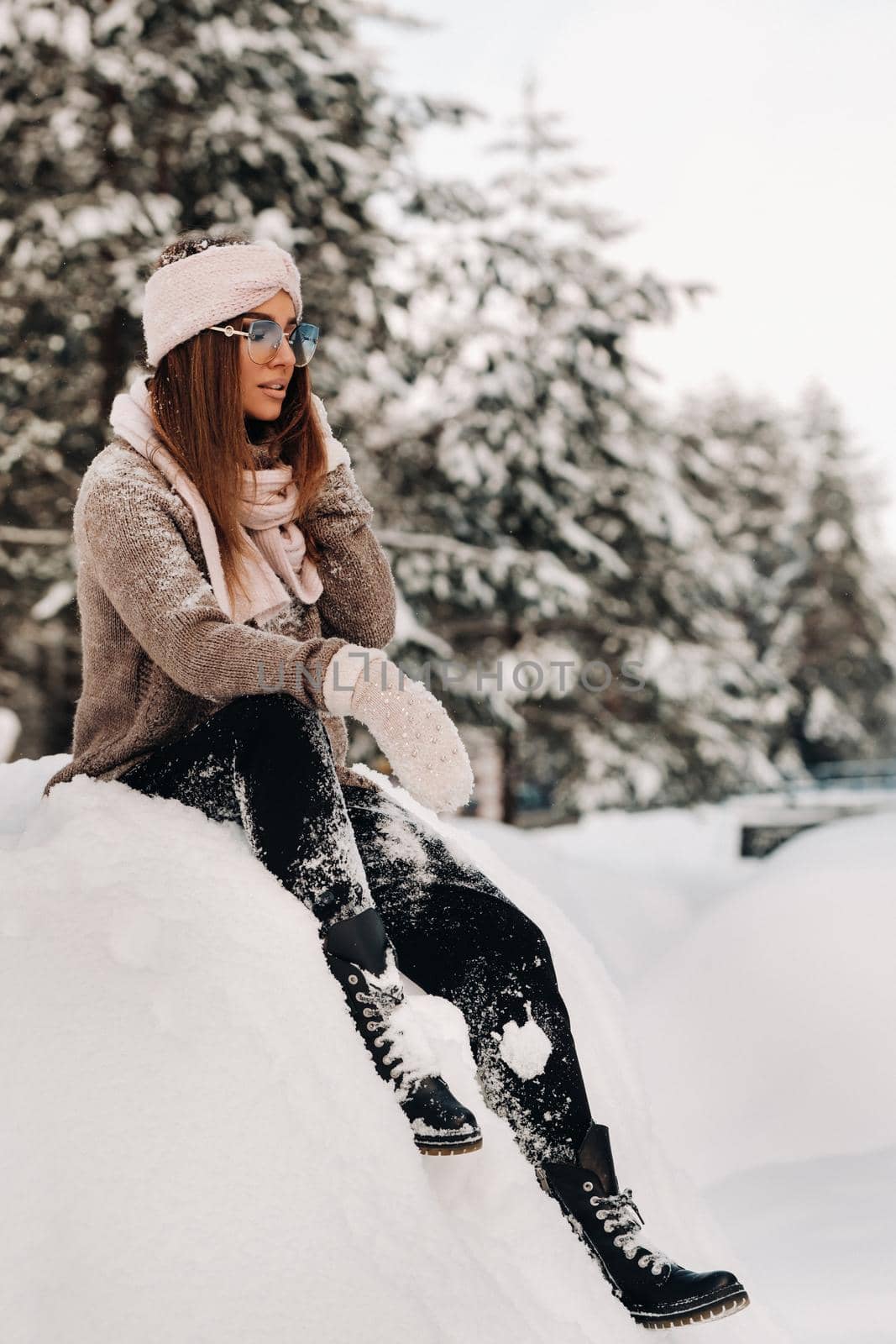 A girl in a sweater and glasses in winter sits on a snow-covered background in the forest.