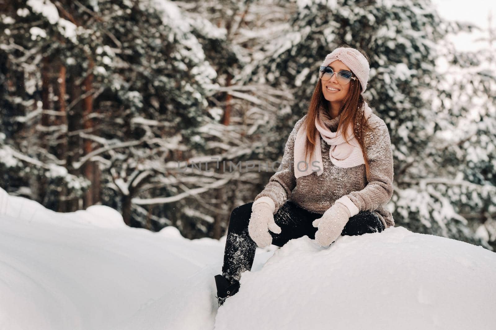 A girl in a sweater and glasses in winter sits on a snow-covered background in the forest.