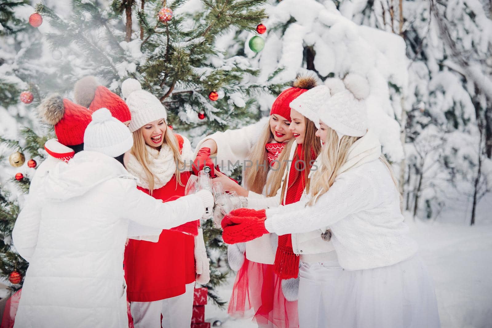 A large group of girls with glasses of champagne in their hands stands in the winter forest.Girls in red and white clothes with new year's drinks in a snow-covered forest. by Lobachad