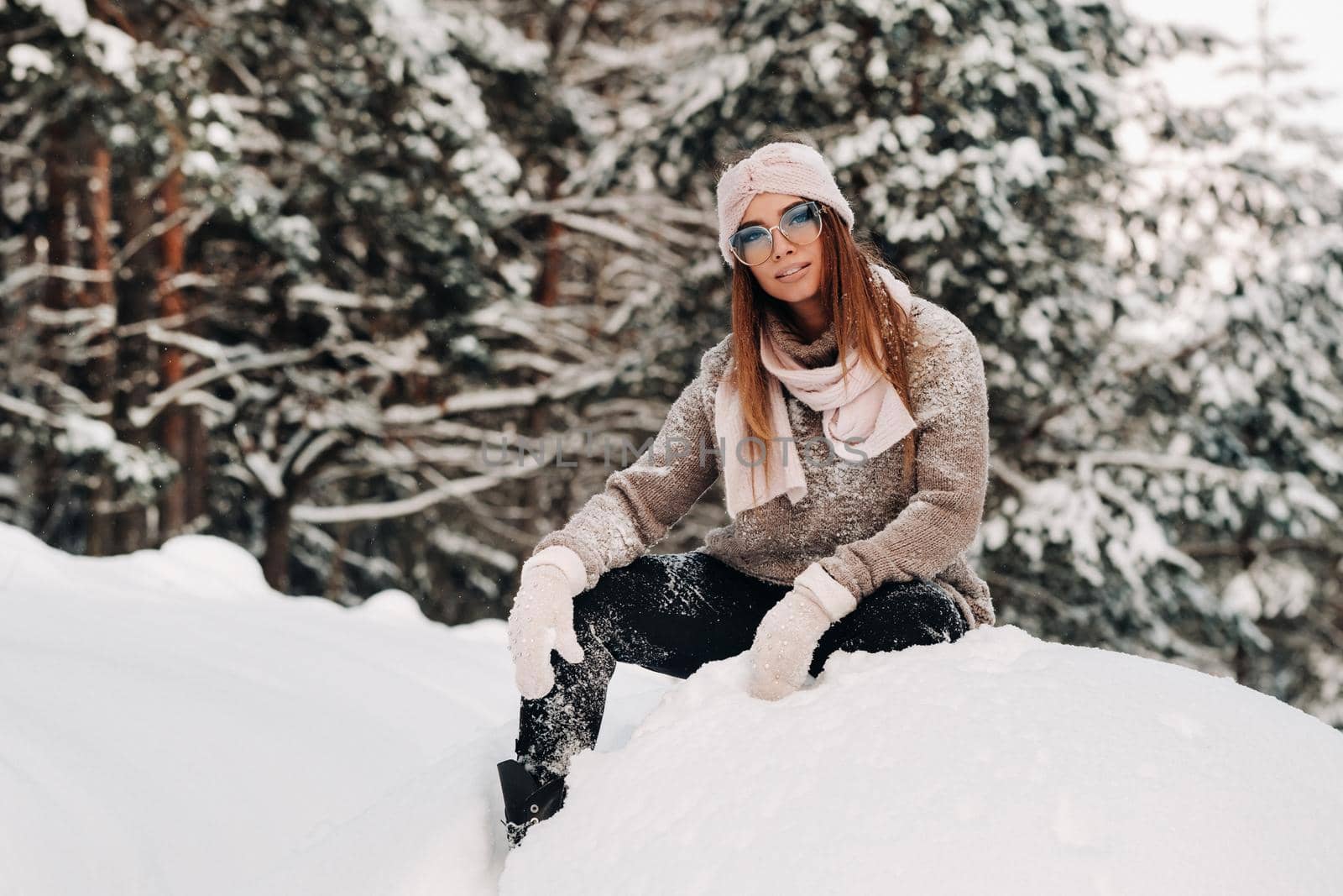 A girl in a sweater and glasses in winter sits on a snow-covered background in the forest.