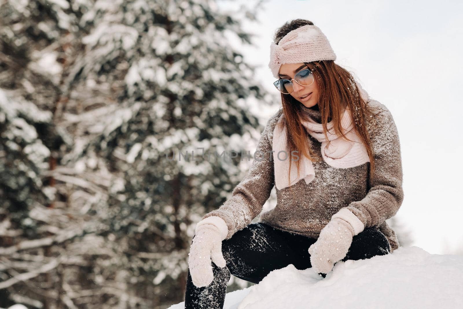 A girl in a sweater and glasses in winter sits on a snow-covered background in the forest.