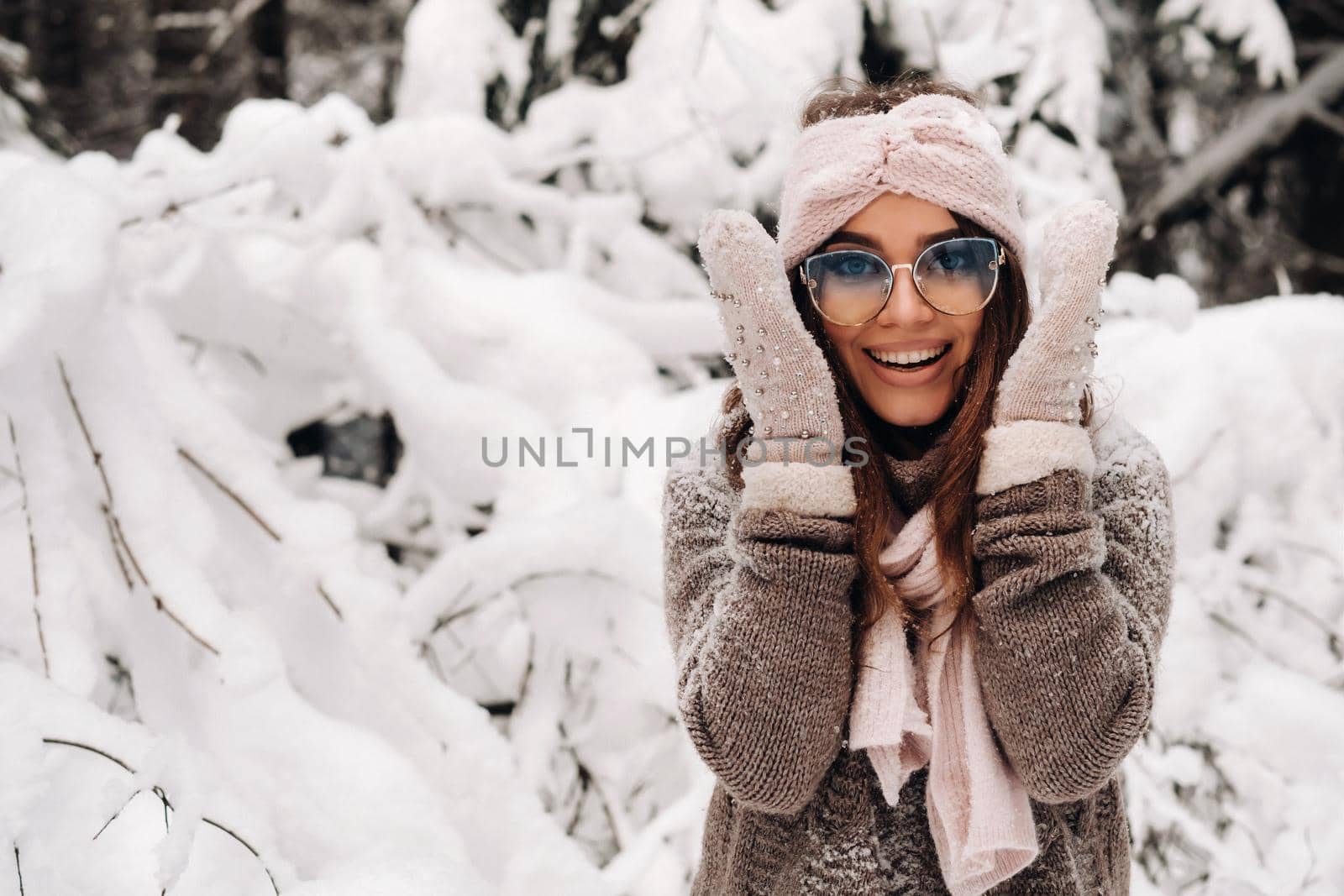 A girl in a sweater and mittens in winter holds her head with her hands in a snowy forest.