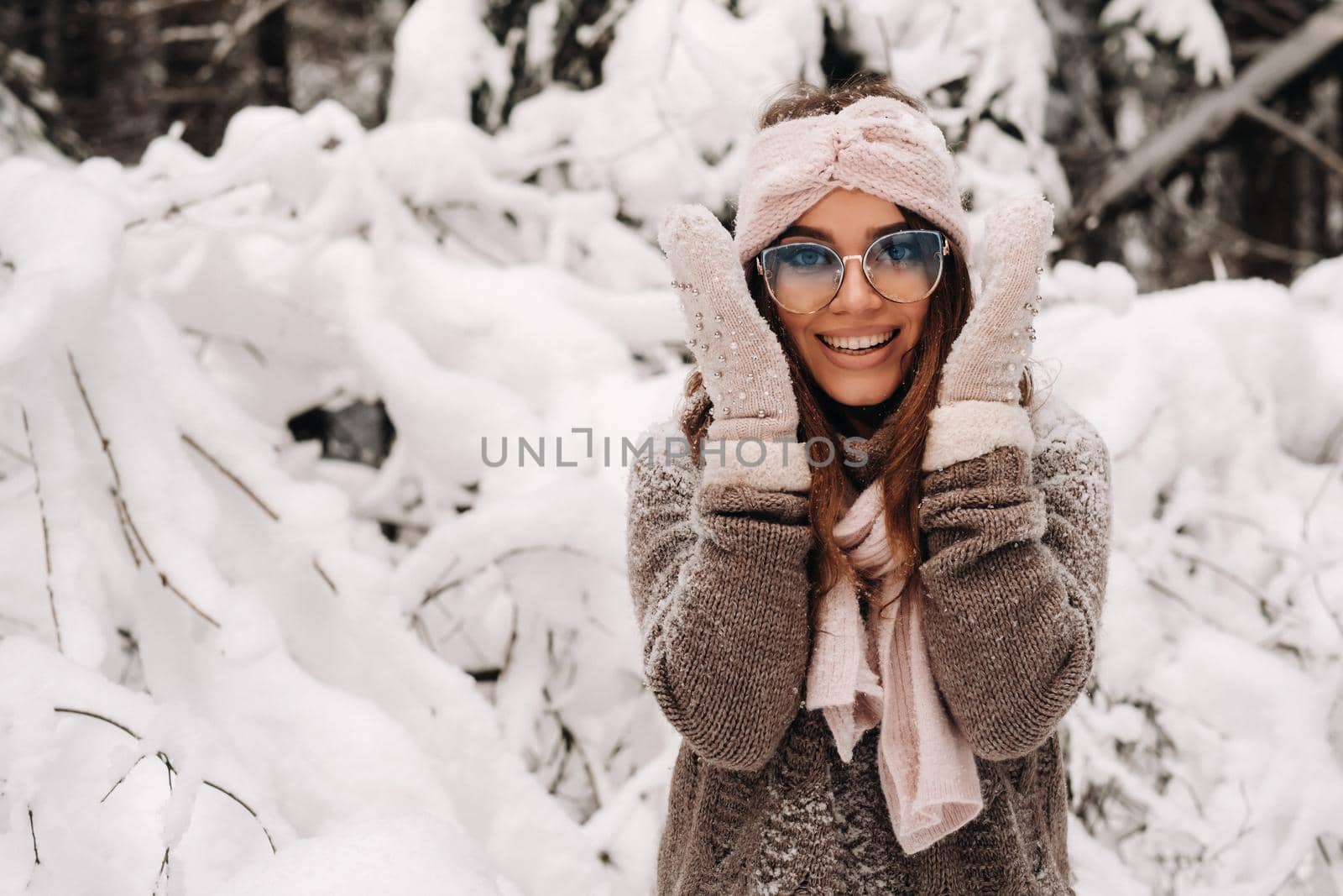 A girl in a sweater and mittens in winter holds her head with her hands in a snowy forest.