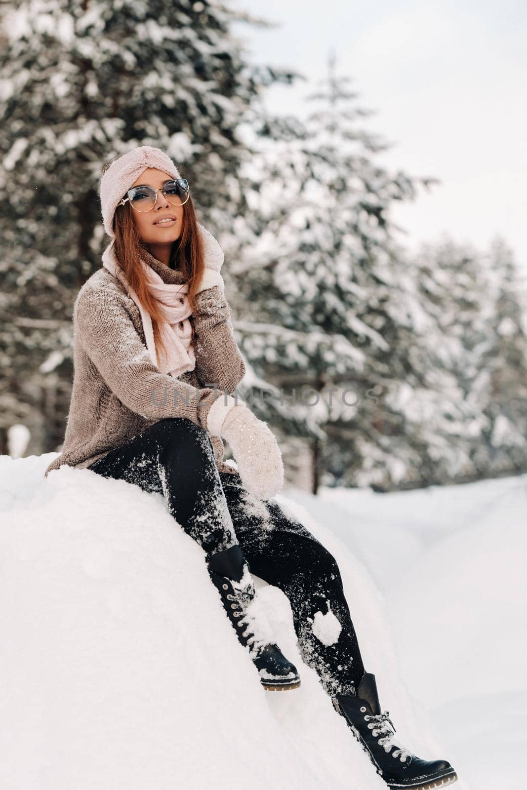 A girl in a sweater and glasses in winter sits on a snow-covered background in the forest.