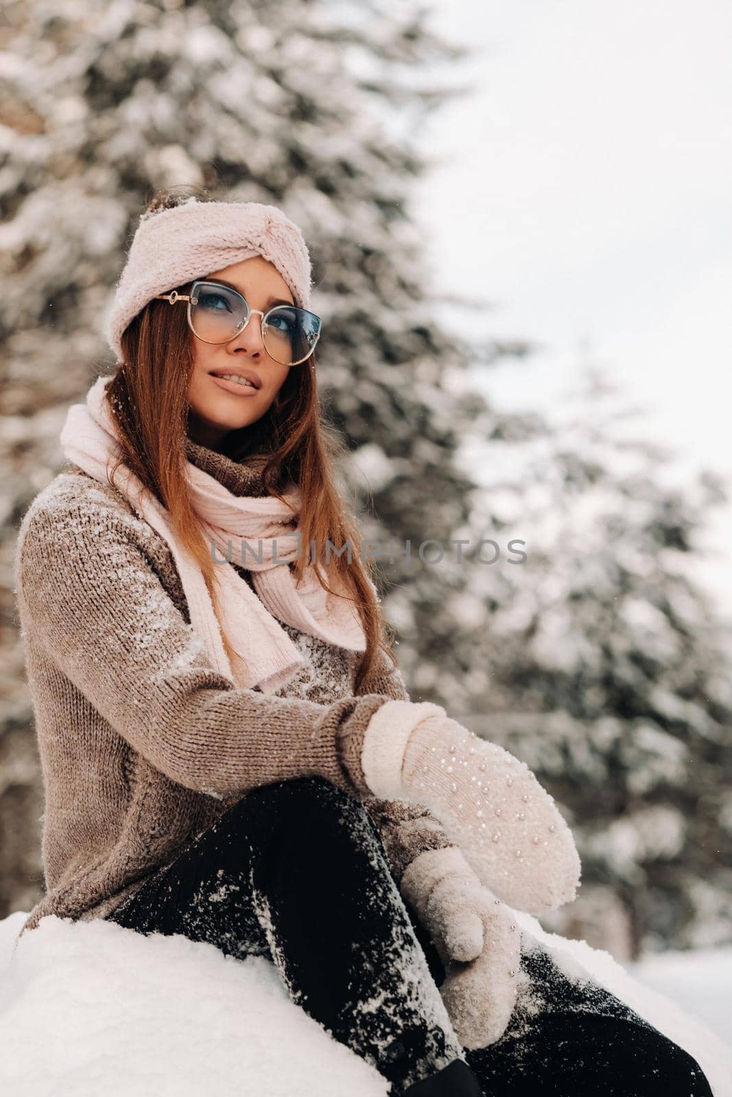 A girl in a sweater and glasses in winter sits on a snow-covered background in the forest.