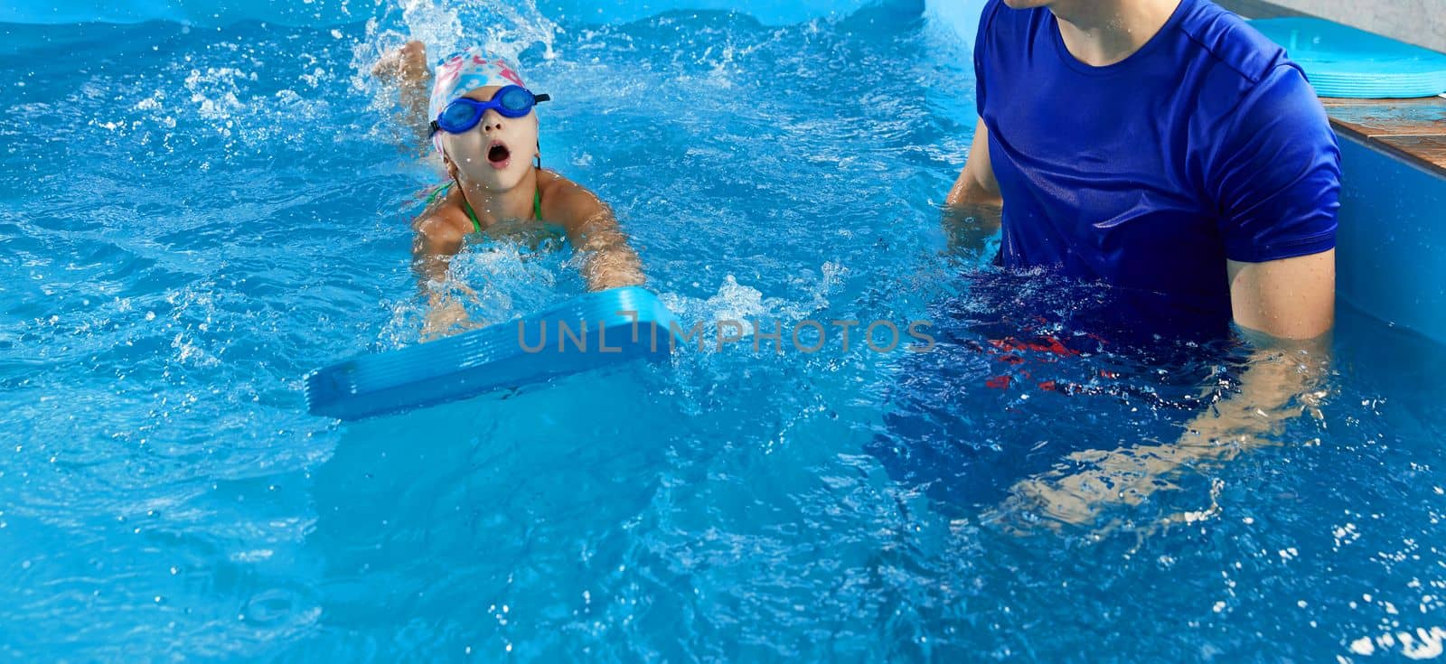 Little girl learning to swim in indoor pool with pool board. Swimming lesson. Active child swims in water with teacher