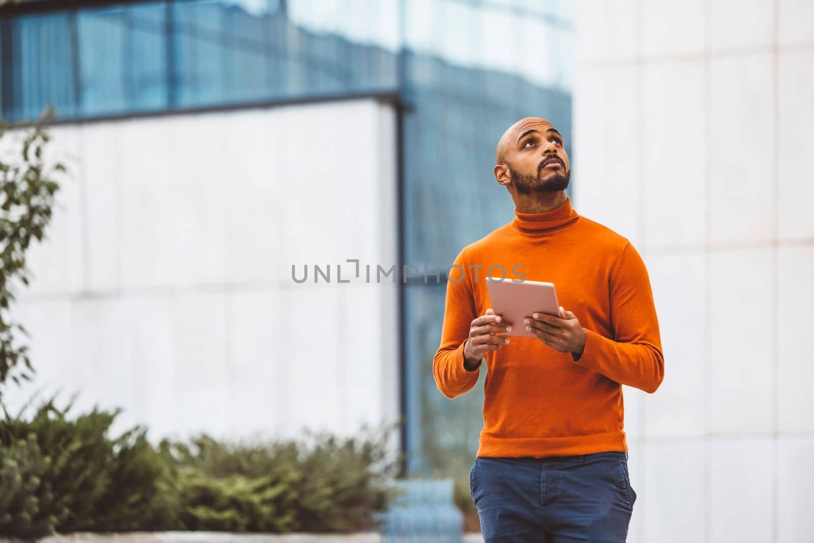 Caucasian man in an orange sweater standing outside in an urban setting holding a digital device, office buildings in the background.