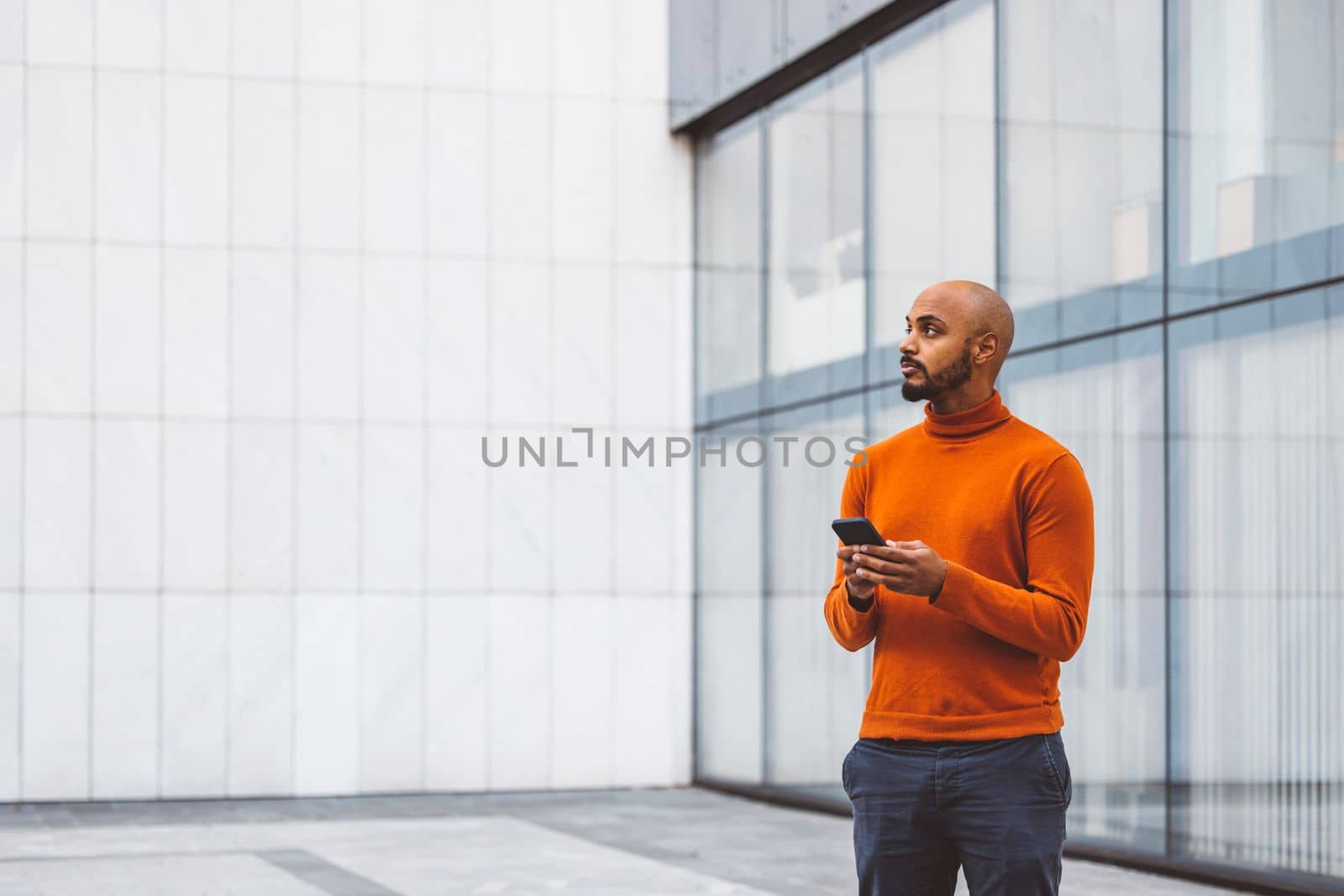 Caucasian man in an orange sweater standing outside in an urban setting holding a digital device, office buildings in the background.