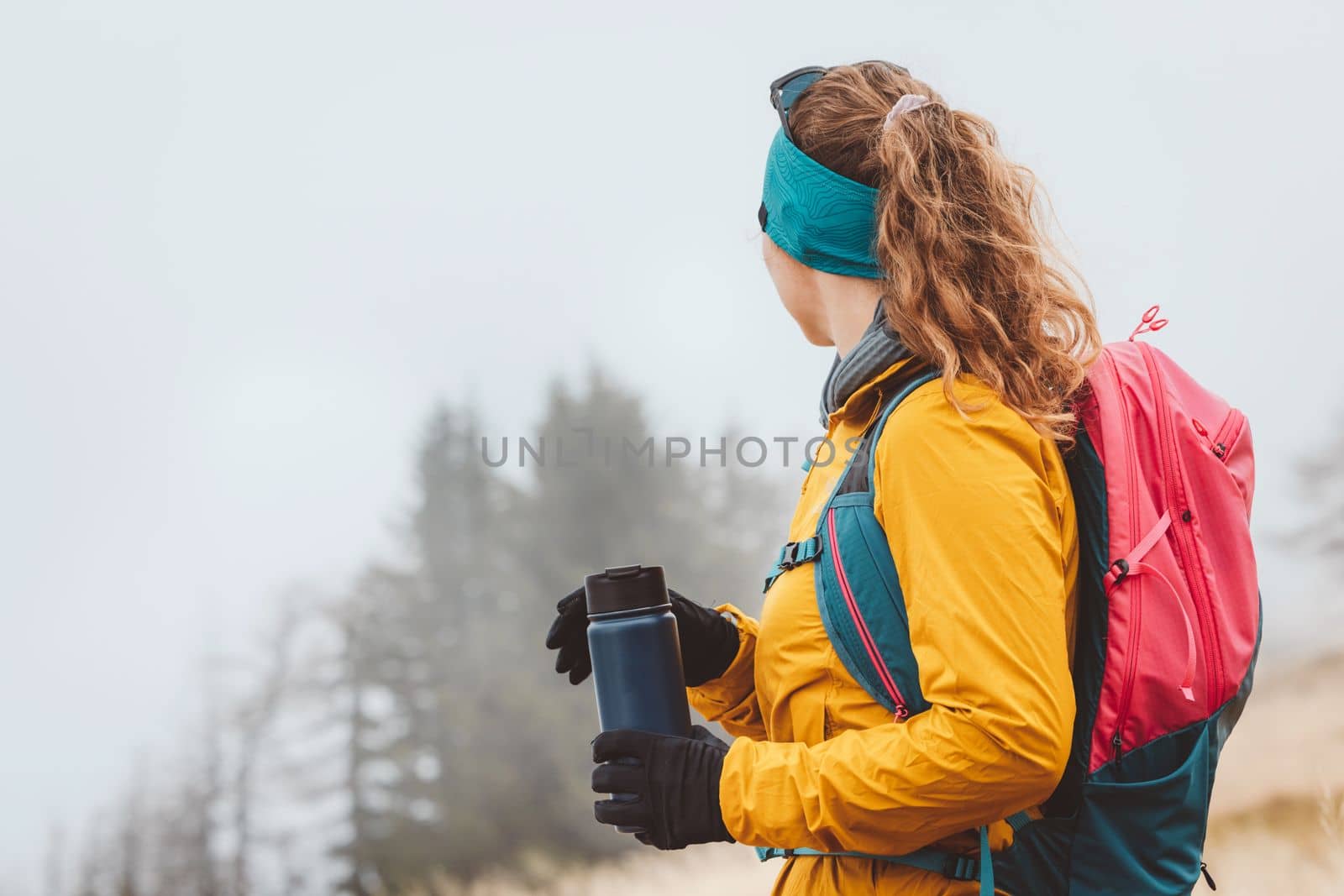 Waist up woman hiker looking away from the camera holding a blue thermo flask in her hands, hiking on a foggy autumn day,