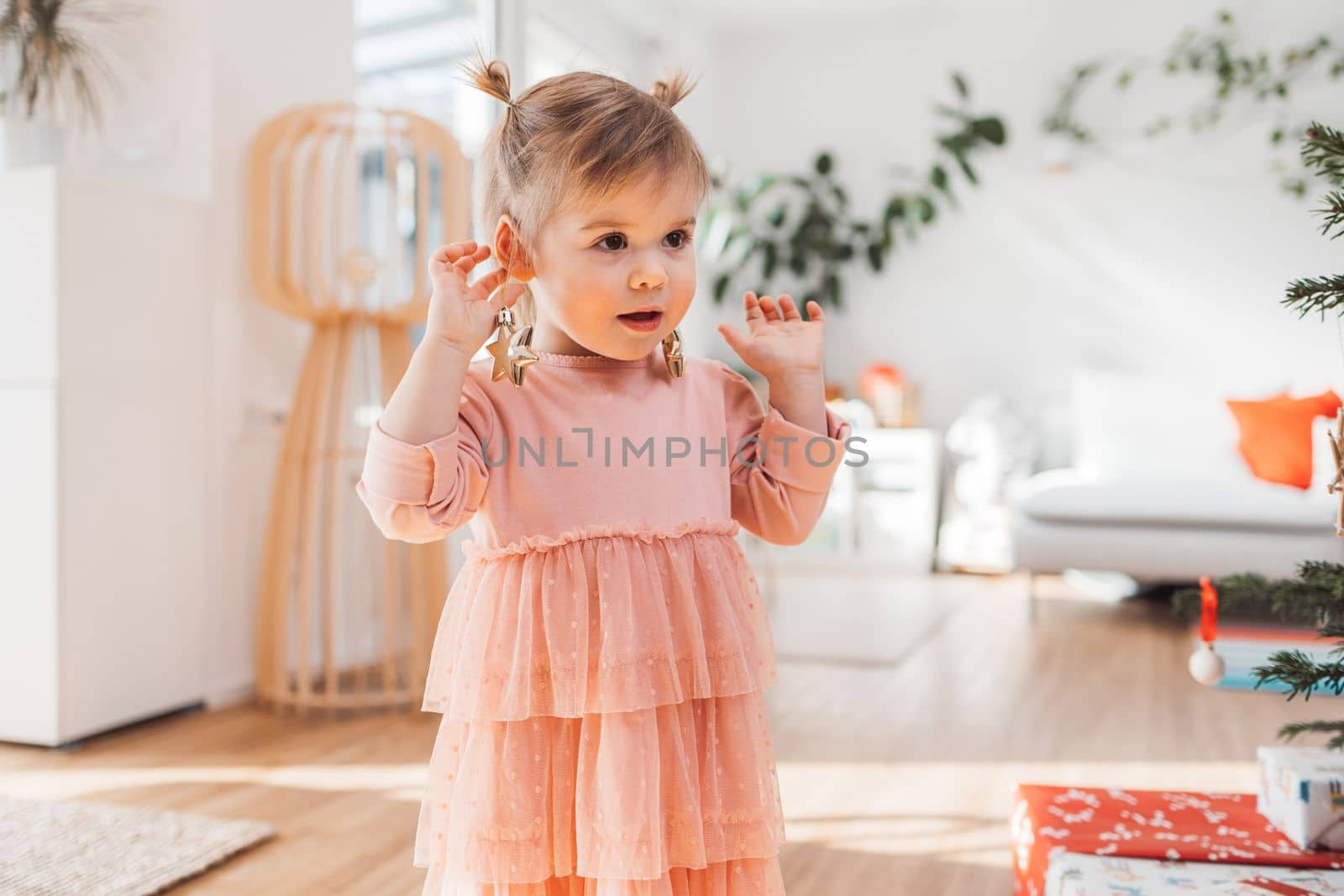 Cheerful young girl, a toddler, enjoying some lollipop while decorating the Christmas tree. Smiling little girl wearing a pink dress and two ponytails.