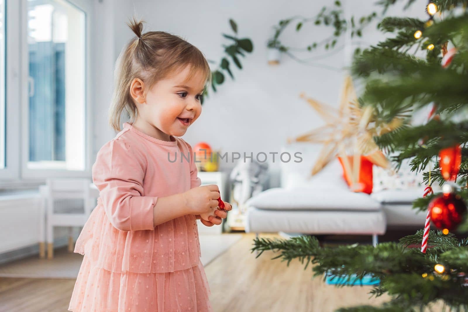 Smiling baby girl in pink dress deciding where to put the ornament she's holding in her arms by VisualProductions