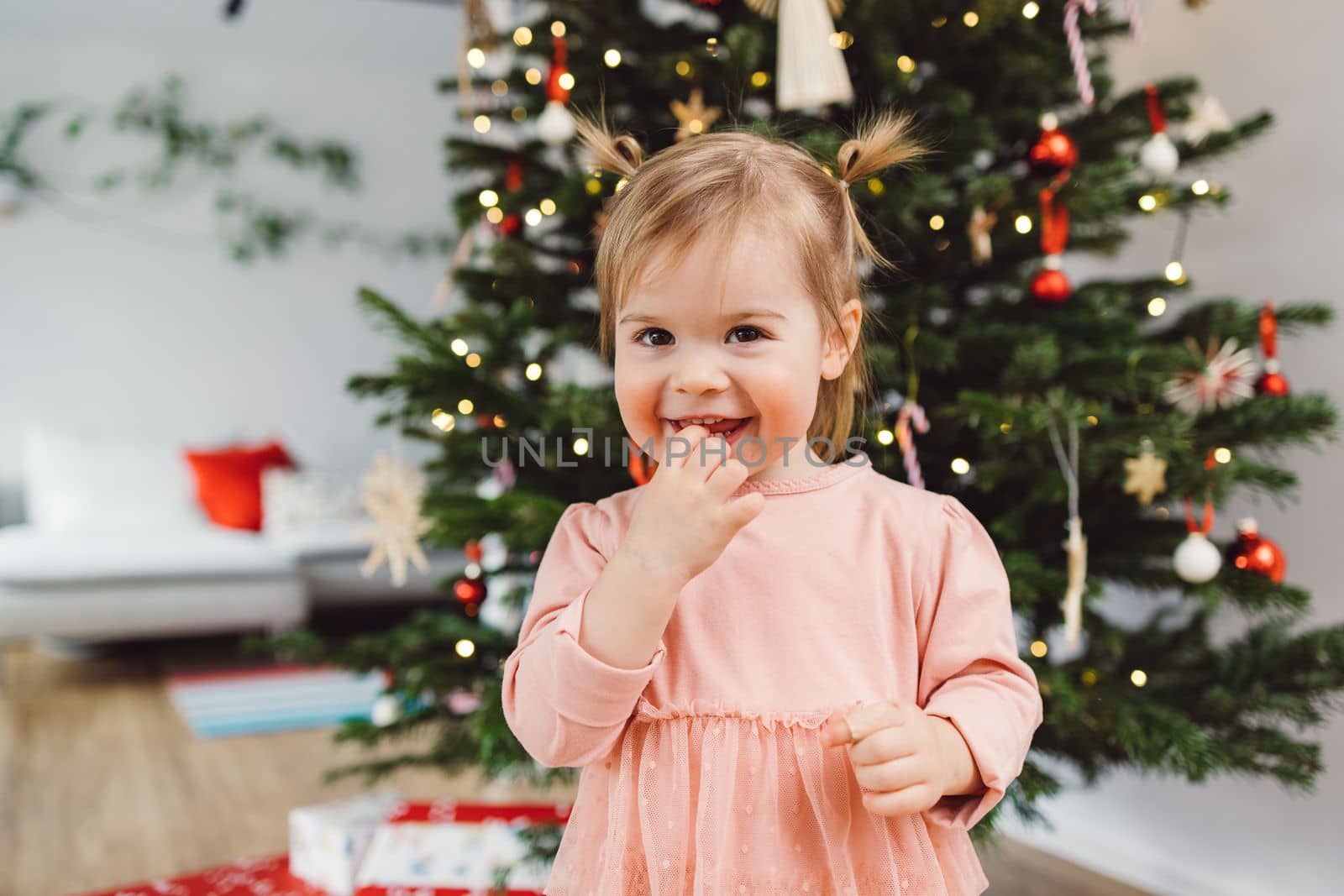 Cute little baby girl smiling at the camera chewing on her fingers, standing in front of the Christmas tree by VisualProductions