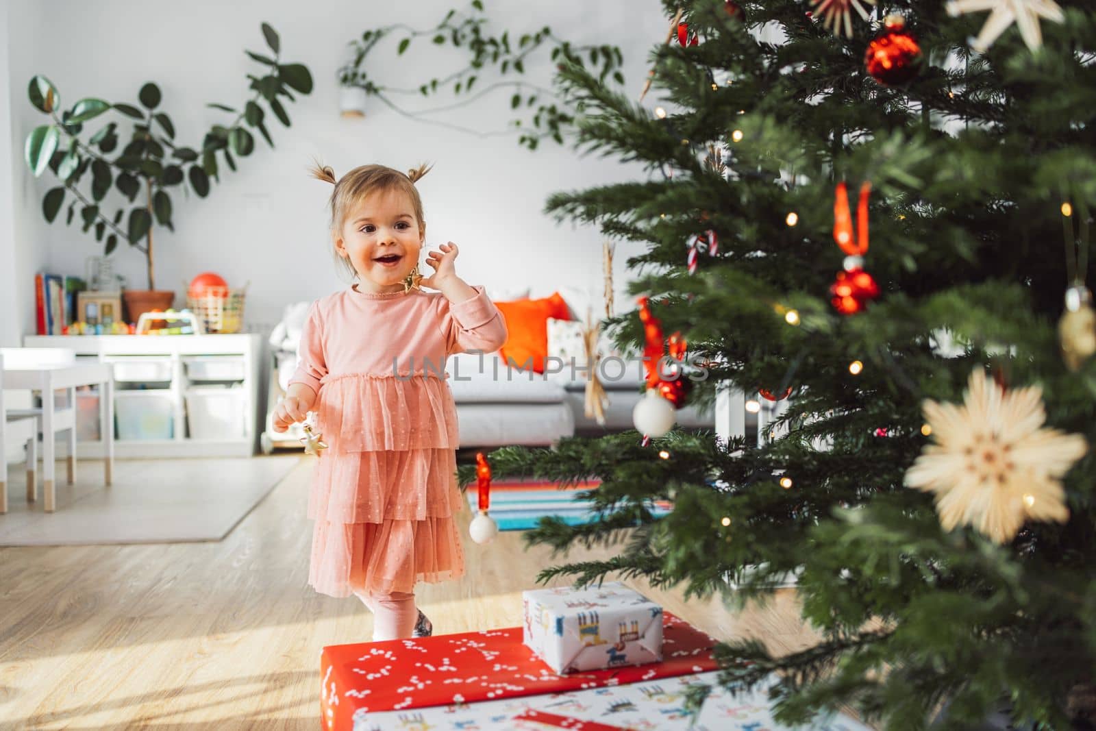 Joyfull little girl playing with Christmas ornament with a big smile on her face, walking past the Christmas tree deciding where to put the ornaments by VisualProductions