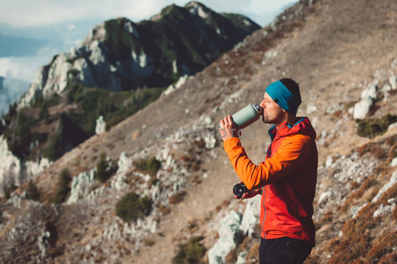 Side view of a man in orange jacket drinking from a thermo flask while looking at the view. Autumn hike.