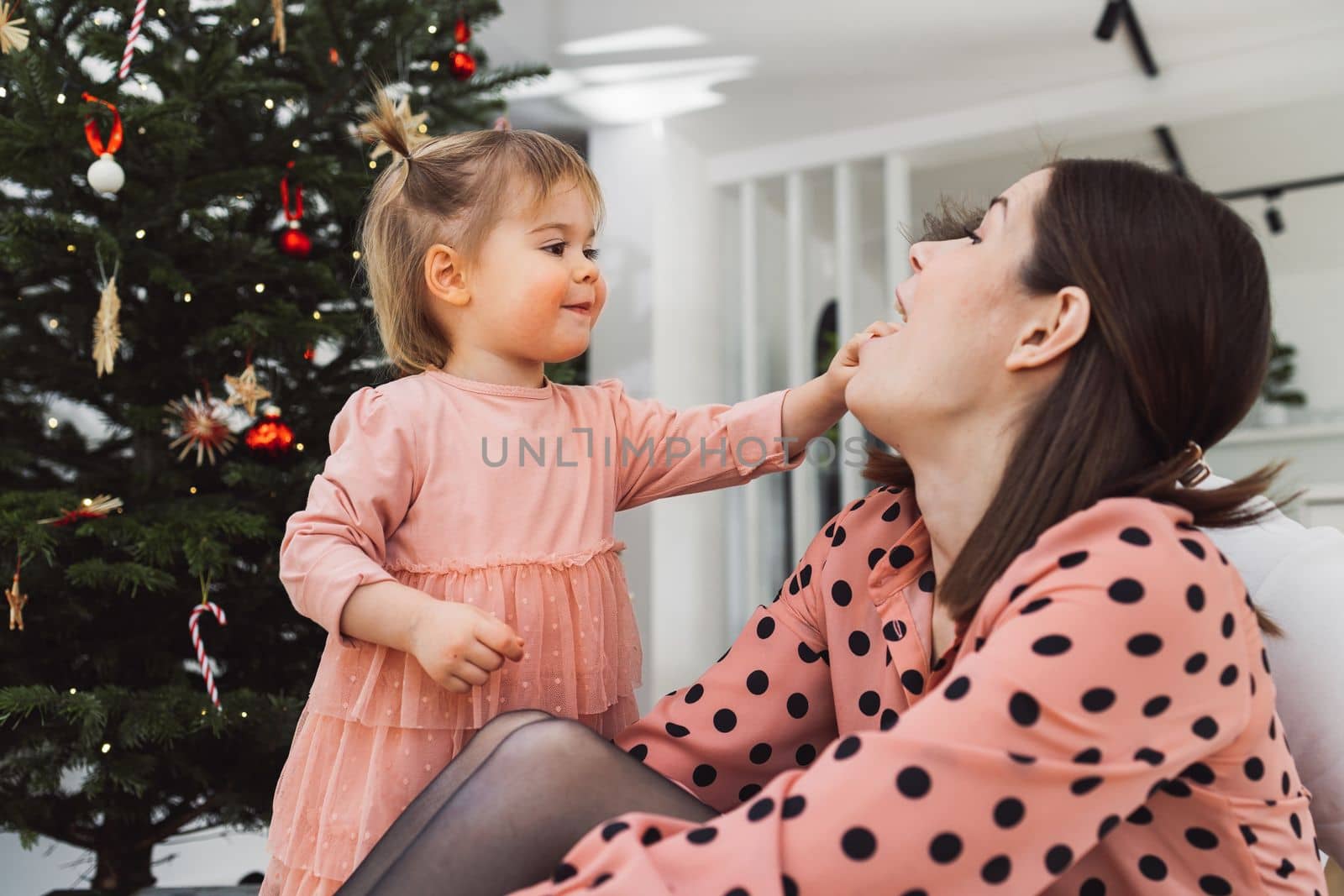 Young caucasian woman, mom, spending time with her toddler, young girl wearing a pink dress matching her mom. Mom and daughter decorating for Christmas together having fun. Mom holding a smiling little girl in her arms.