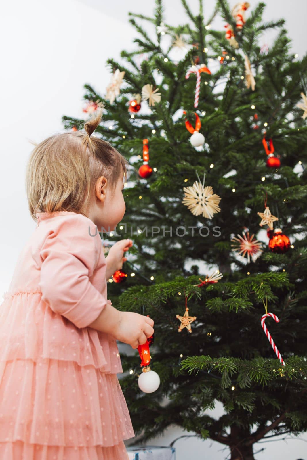 Low angle view, baby girl in pink dress looking up at the Christmas tree, deciding where to put the ornament by VisualProductions