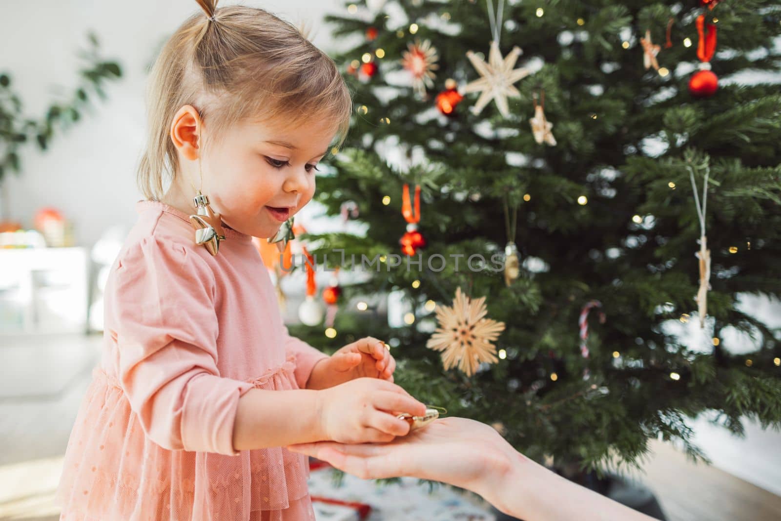 Baby girl in pink dress with Christmas ornaments hanging off her ears, taking another ornament from moms hand by VisualProductions