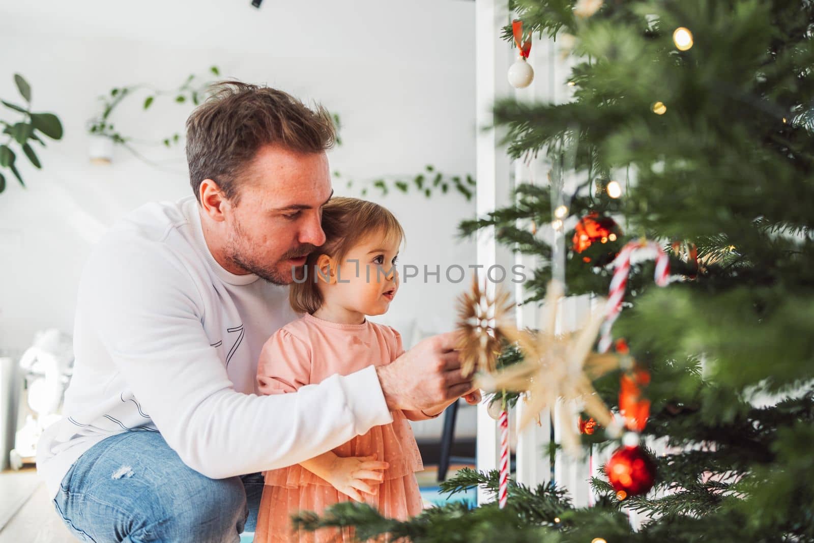 Portrait of young caucasian man with his daughter, decorating the Christmas tree. Loving dad having fun with his little girl on Christmas.