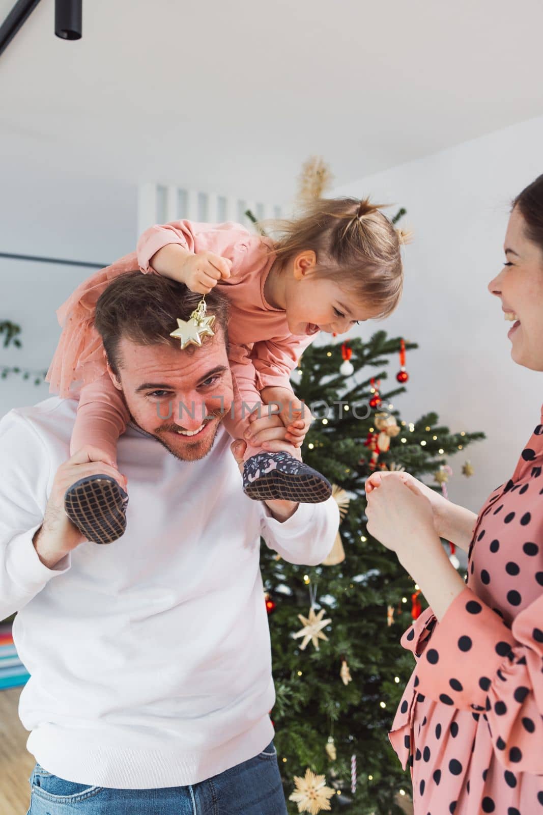 Dad carrying little girl on his shoulders smiling, while mom stands at the side looking at them - Family having fun on Christmas by VisualProductions