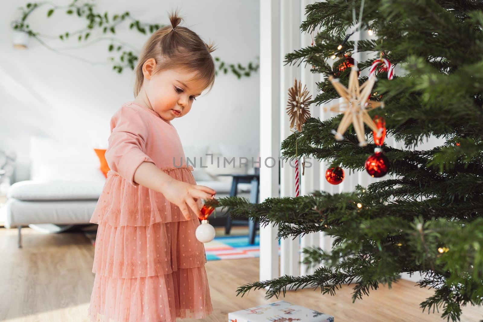 Cheerful young girl, a toddler, enjoying some lollipop while decorating the Christmas tree. Smiling little girl wearing a pink dress and two ponytails.