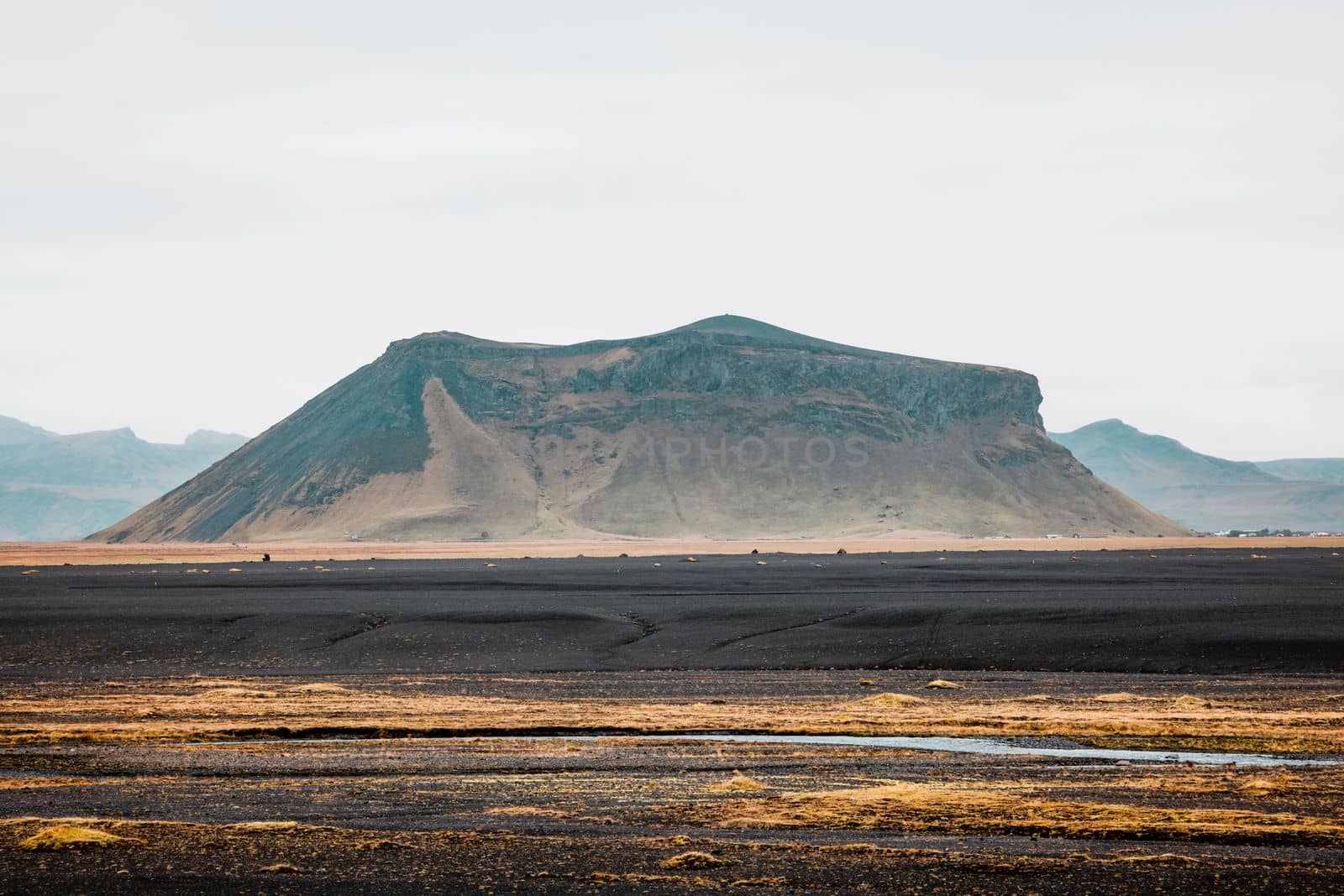 Black volcanic sand beaches in Iceland, a view towards the mainland with a river running trough and some yellow foliage around the water. A mountain right in front.