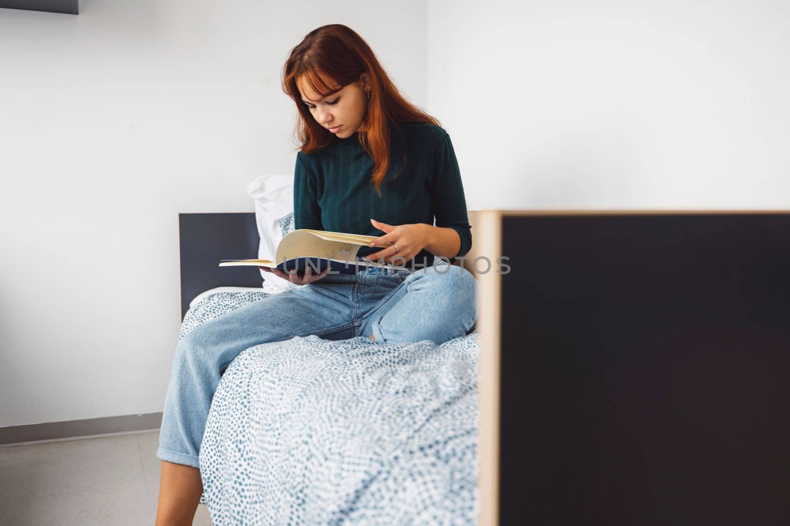 Young caucasian woman, college student studying in her dorm room, sitting on the bed. Bright room with lots of natural light.