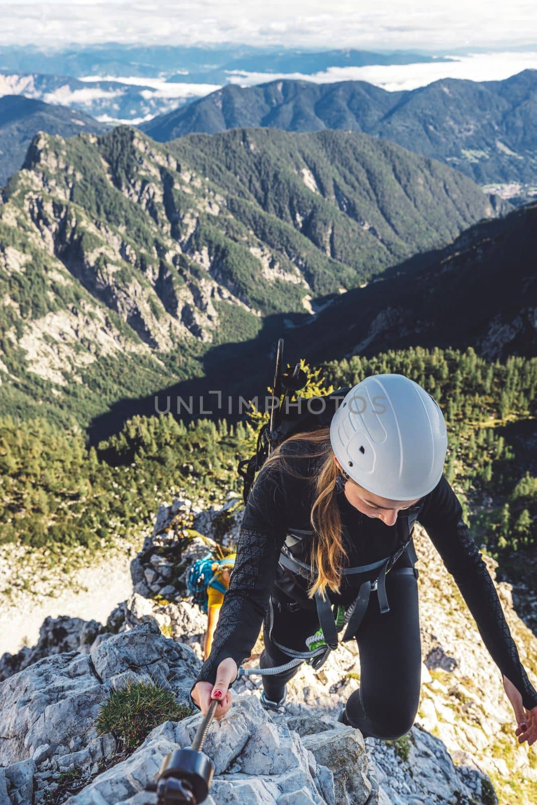 Caucasian woman mountaineer climbing up the mountain on via ferrata trail, high up in the mountain peaks. Woman wearing a protective head gear and uses a safety harness while she climbs.