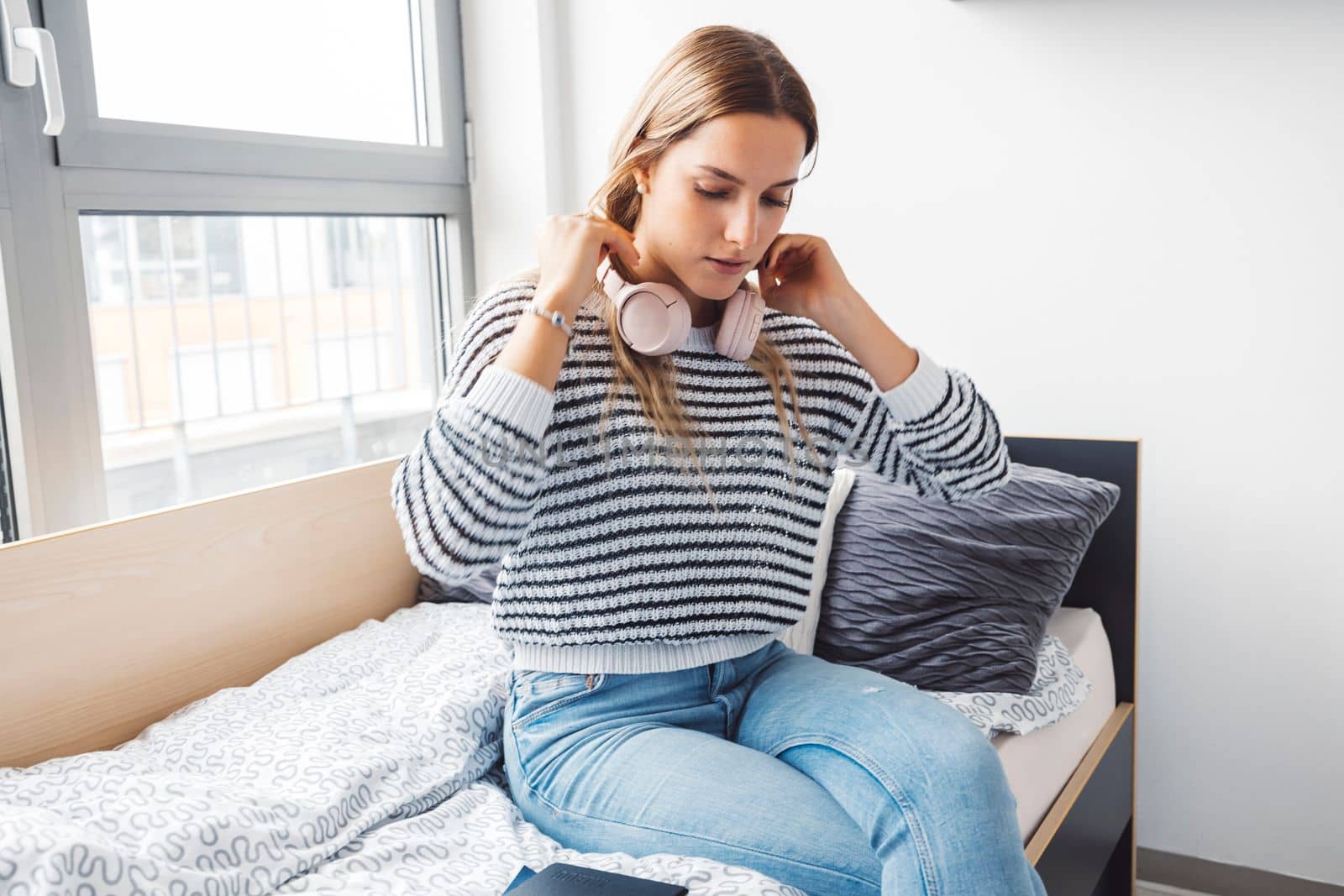 Blonde young caucasian woman lying on her dorm room bed next to a window putting on headphones, listening to music on a cold cloudy autumn day.