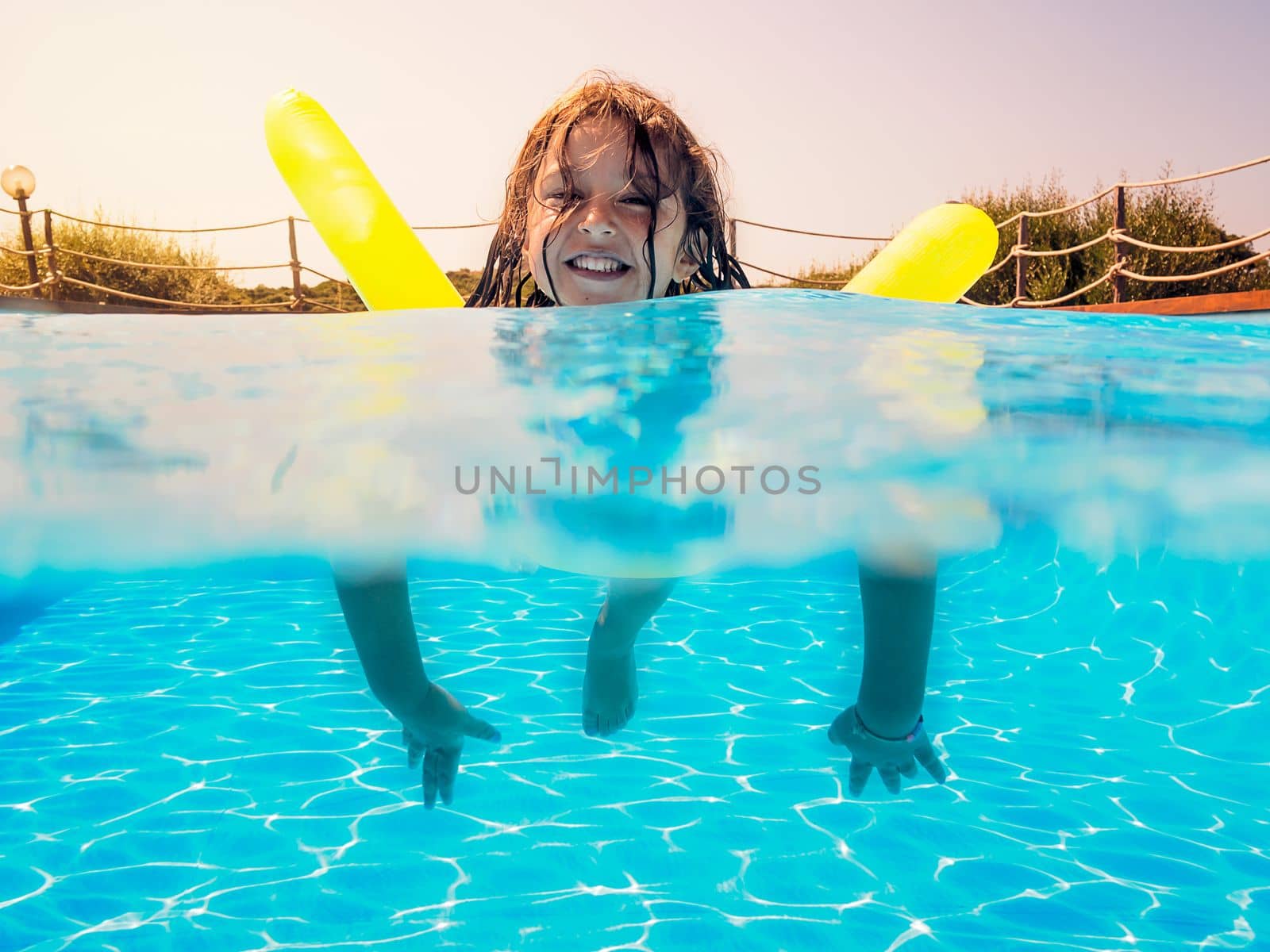 Underwater view of a girl swimming smiling toward the camera with her yellow float in the pool, behind her the glow of the summery sun gleams golden