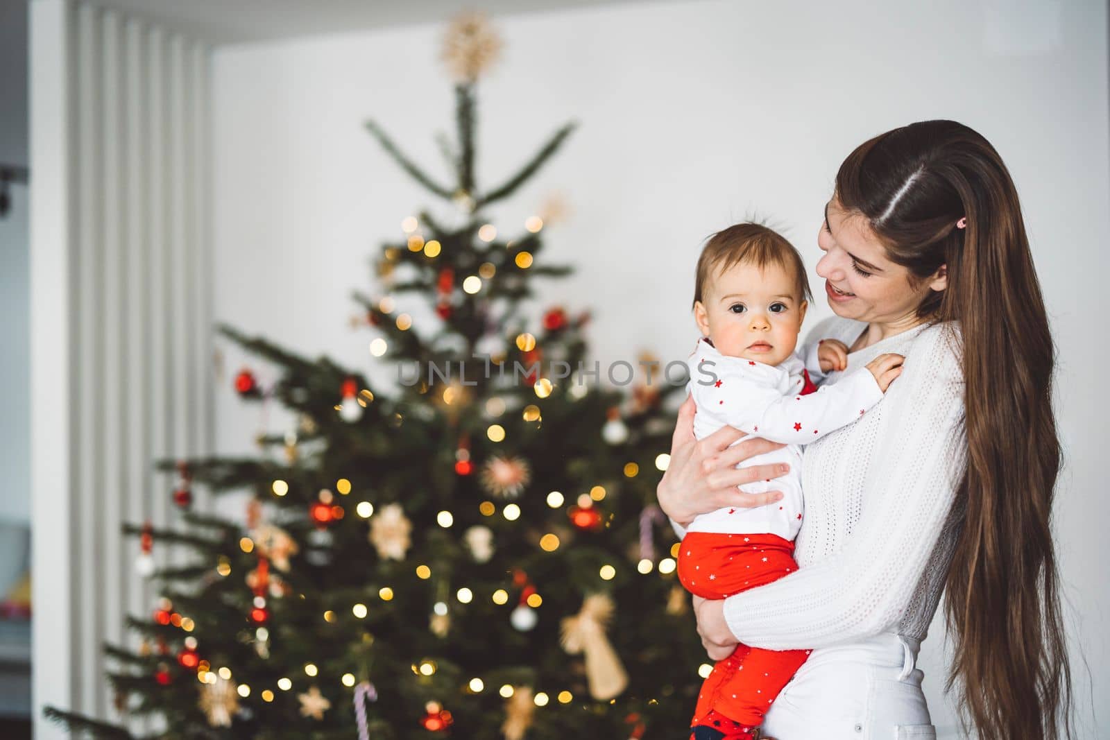 Portrait of young mom and her daughter, baby girl having fun on Christmas, decorating the Christmas tree in festive outfits. Smiling baby girl playing with her mom.