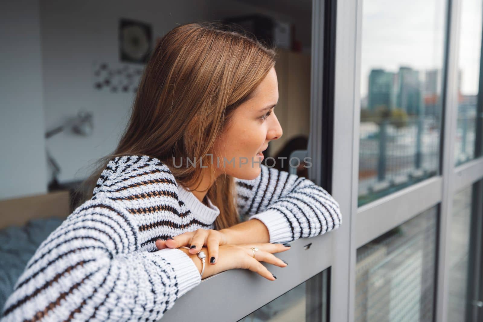 Young blonde caucasian woman looking out the window of her dorm room getting some fresh air on a cold autumn day.