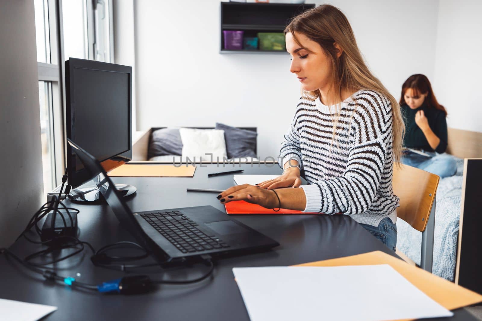 Young caucasian woman, college student studying in her dorm room, sitting by the desk. Bright room with lots of natural light.