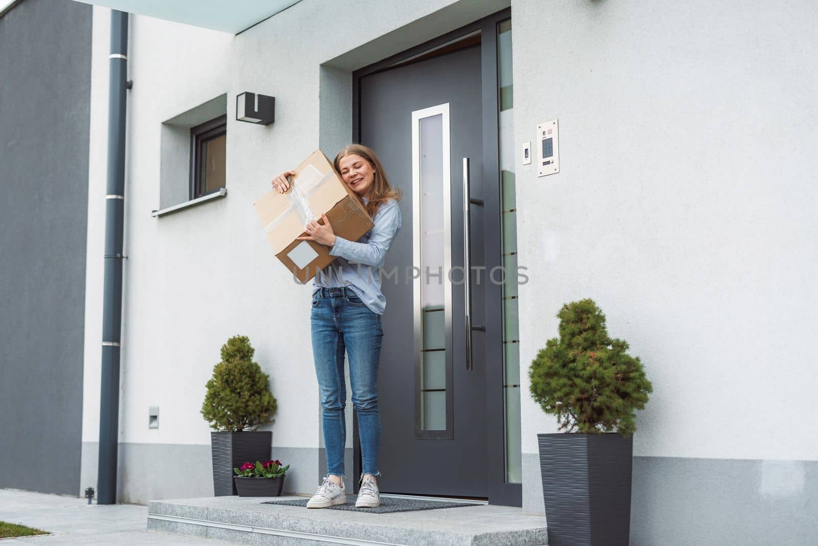 Thrilled caucasian woman standing at the front door with a big cardboard box that just came in the mail. Woman receiving an exciting package, holding it in her arms.