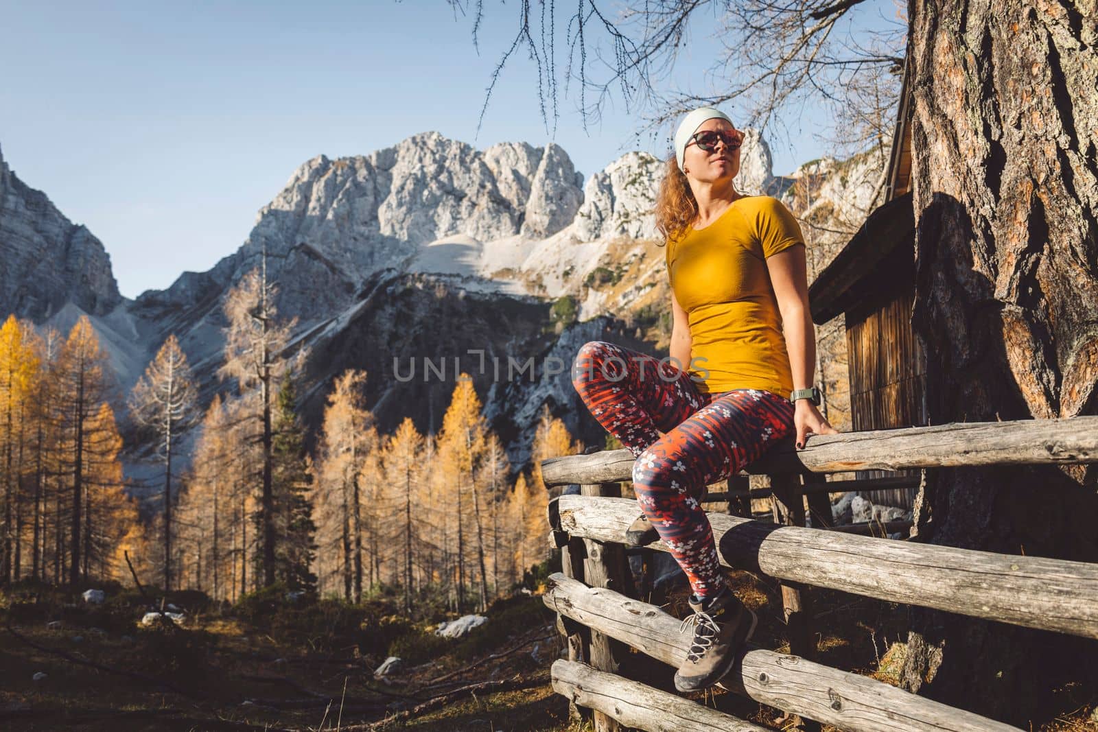 Side view of young caucasian woman hiker in colorful clothes, hiking gear, sitting on a wooden fence somewhere up in the mountains, enjoying the view of the mountain peaks on a beautiful sunny autumn day.