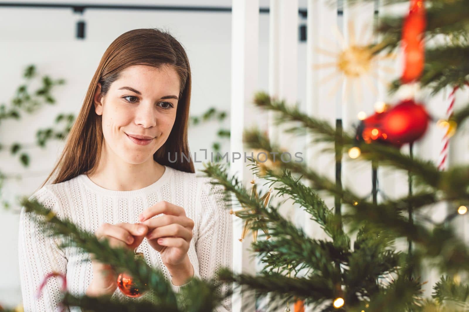 Beautiful smiling cheerful caucasian woman dressed in white outfit decorating a natural Christmas tree, putting red ornaments on the tree, holding a red Christmas ornament.