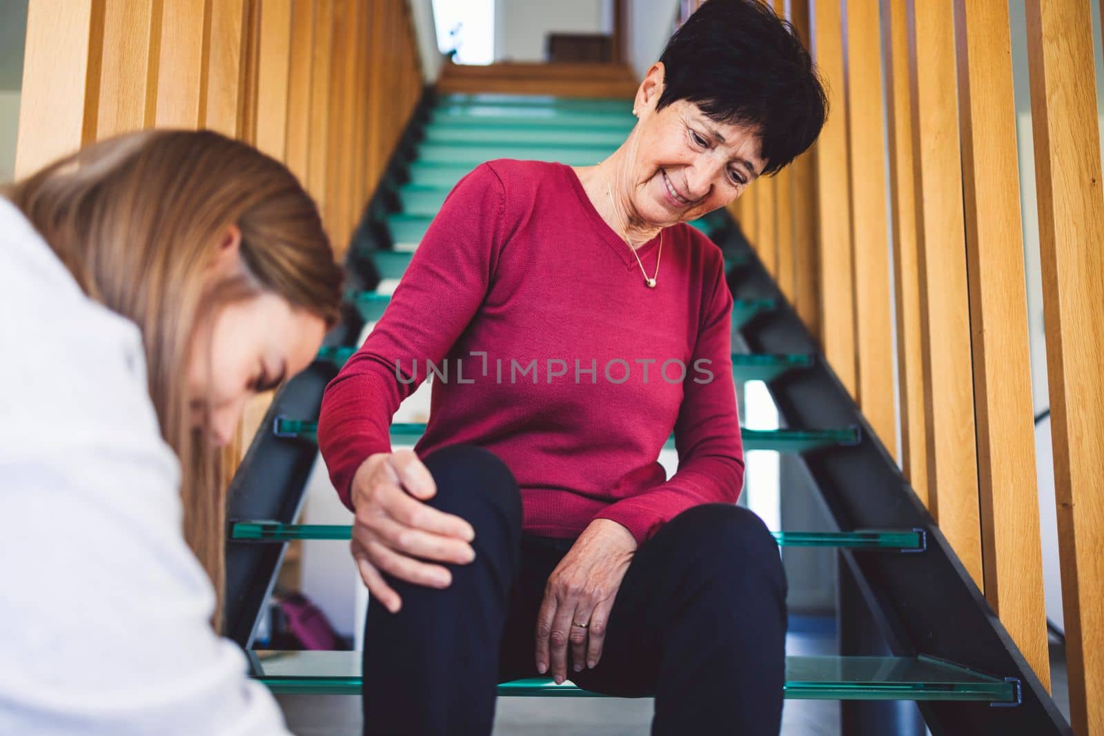 Senior woman sitting on the stairs patiently waiting for nurse to tie up her shoes by VisualProductions