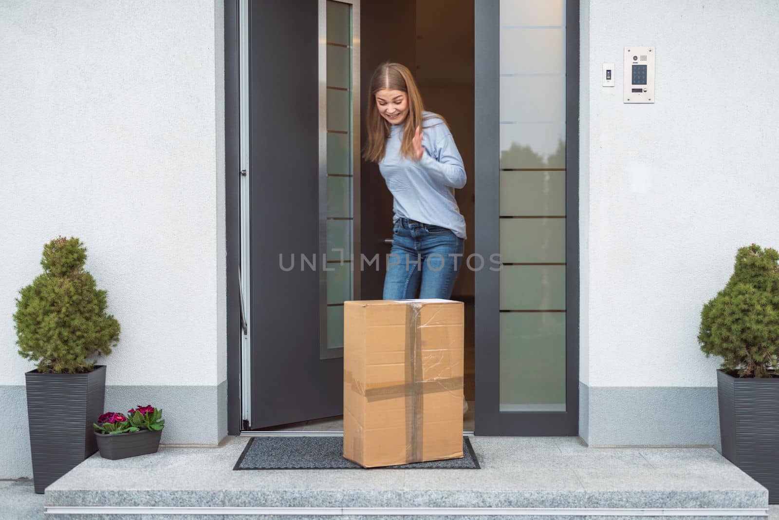 Thrilled caucasian woman standing at the front door with a big cardboard box on the doormat that just came in the mail. Woman receiving an exciting package.