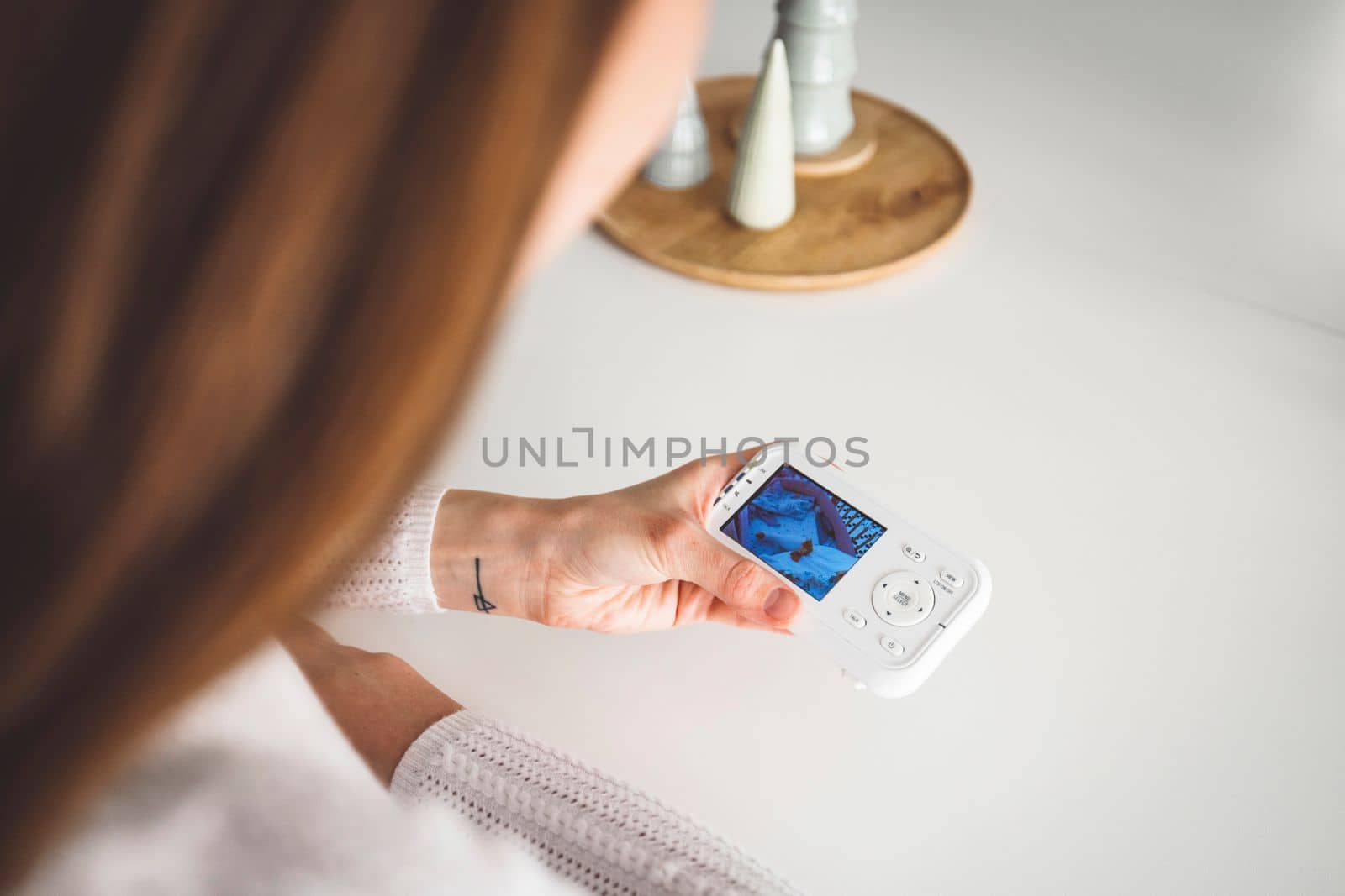 Young caucasian woman dressed in white sitting by the white kitchen island looking at the baby monitor, watching her baby sleep.