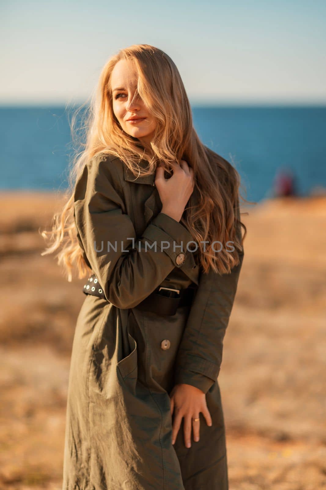 Portrait blonde sea cape. Calm young blonde in a khaki raincoat stands on the seashore.