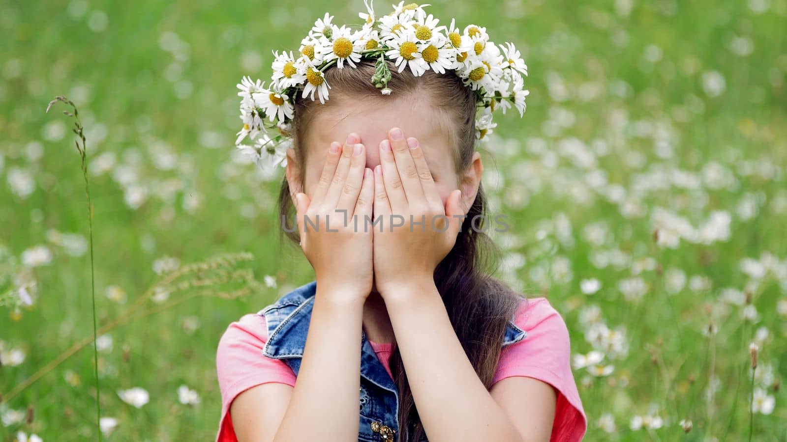 On a chamomile lawn, a sweet girl in a wreath of daisies, smiling, pressing her hands to her cheeks. High quality photo