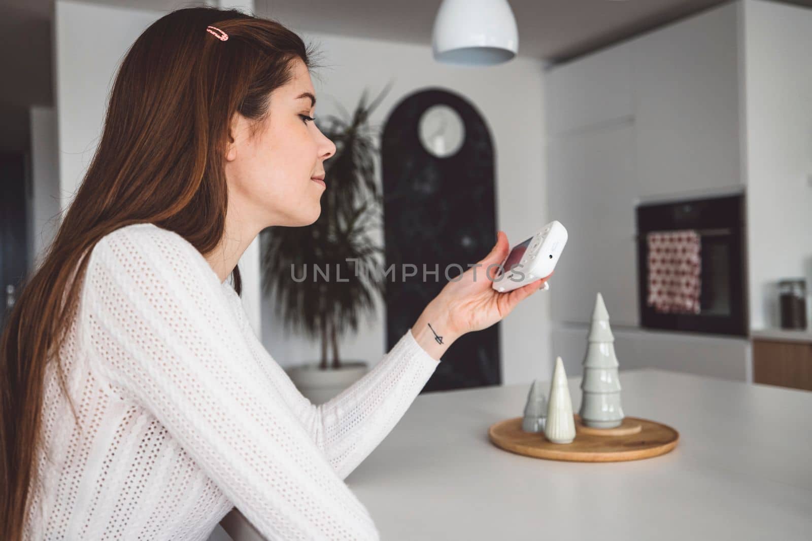 Young caucasian woman dressed in white sitting by the white kitchen island looking at the baby monitor, watching her baby sleep.