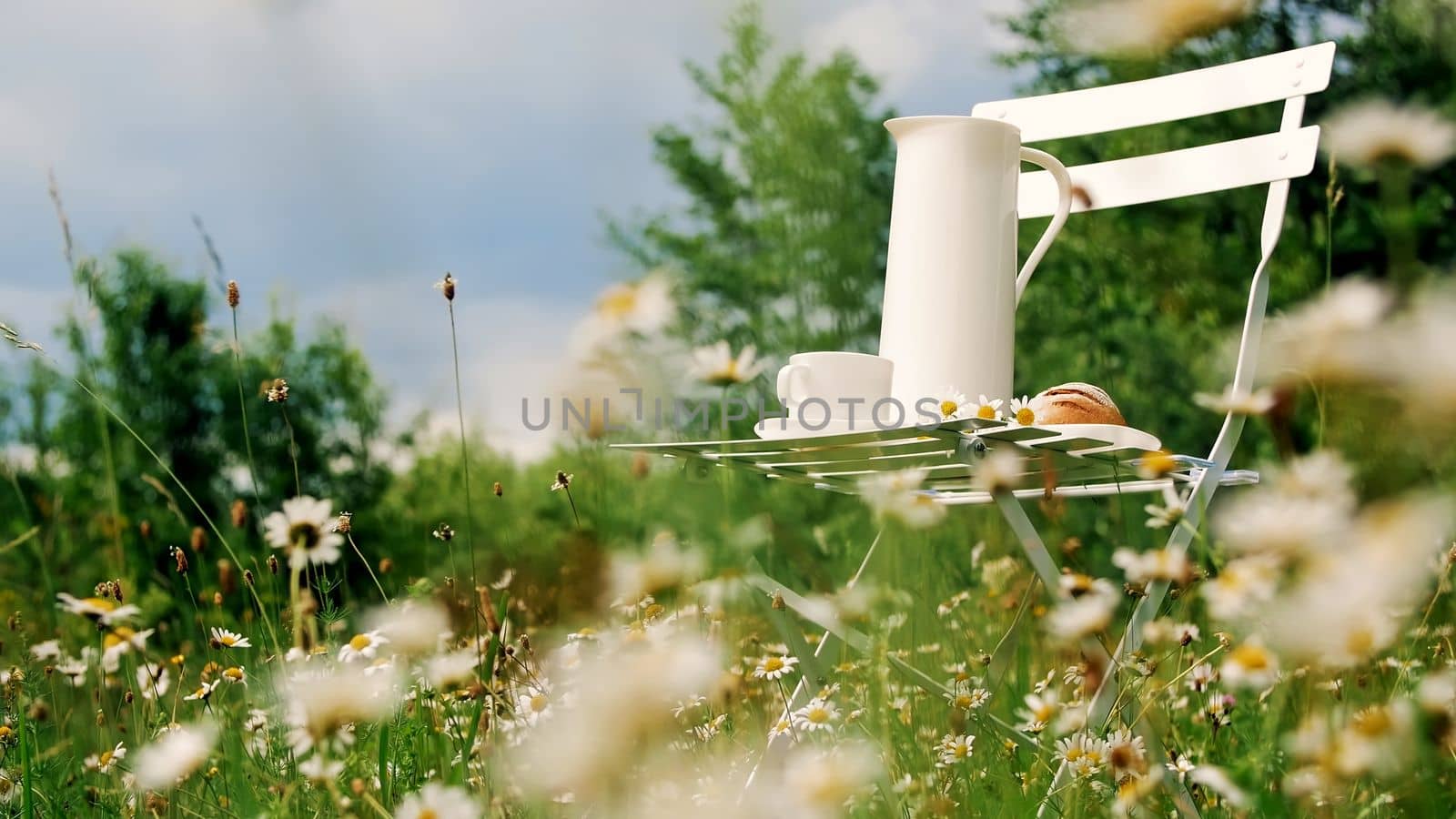 Among the chamomile lawn, against the blue sky is a white chair. On it a composition from a white jug, a white cup with tea, a batch and a bouquet of chamomiles. High quality photo