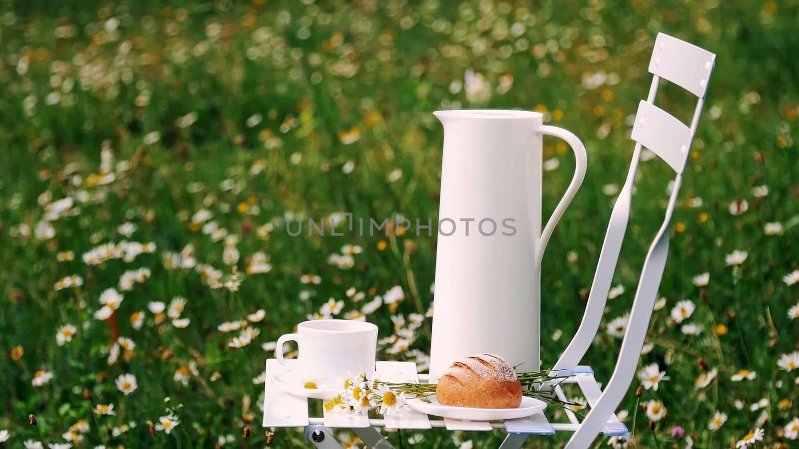 Among the chamomile lawn stands a white chair. On it there is a composition of a white jug, a white cup with tea, a bread and a bouquet of chamomiles. High quality photo