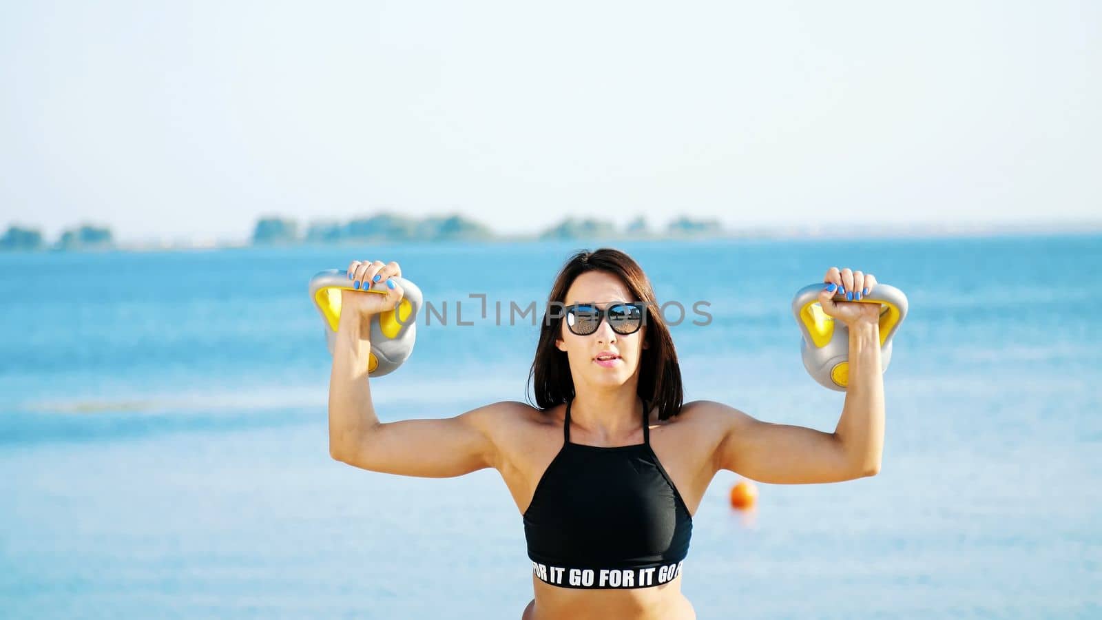 Beautiful, athletic, sexy young woman, in sunglasses, in swimsuit, coach, instructor, performs exercises with weights. On the beach, near the sea, river, in summer, in sun rays. by djtreneryay