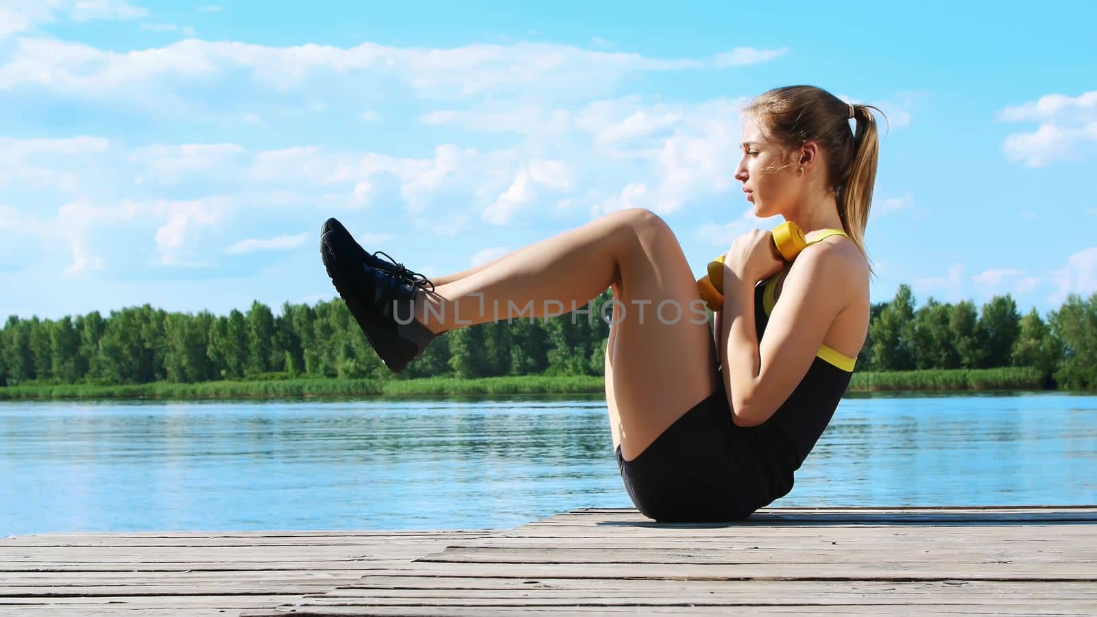 Beautiful, athletic young blond woman stretching, swings press. Lake, river, blue sky and forest in the background, summer sunny day by djtreneryay